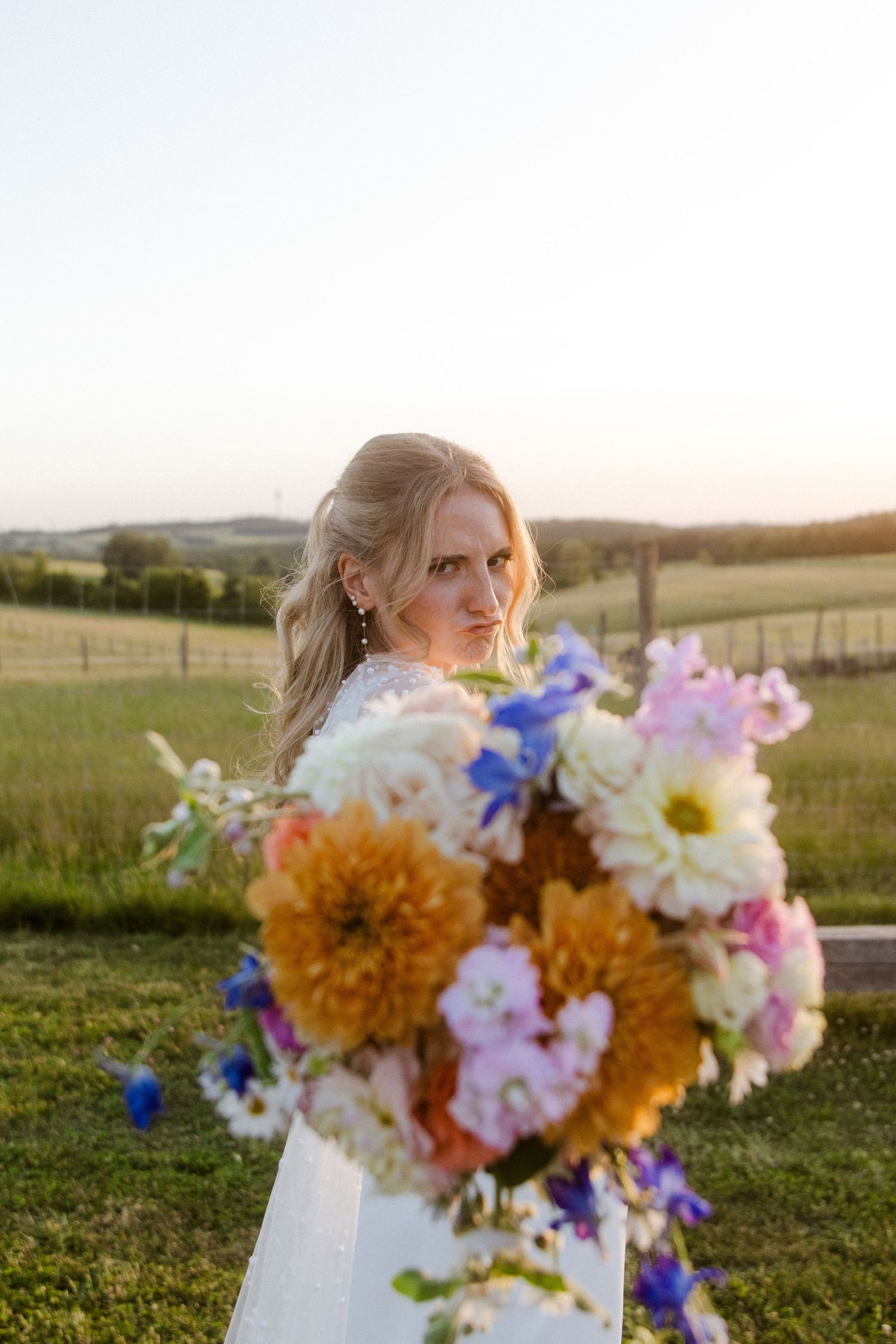 Bride holding a colorful bouquet of dahlias and wildflowers in a sunlit countryside setting.