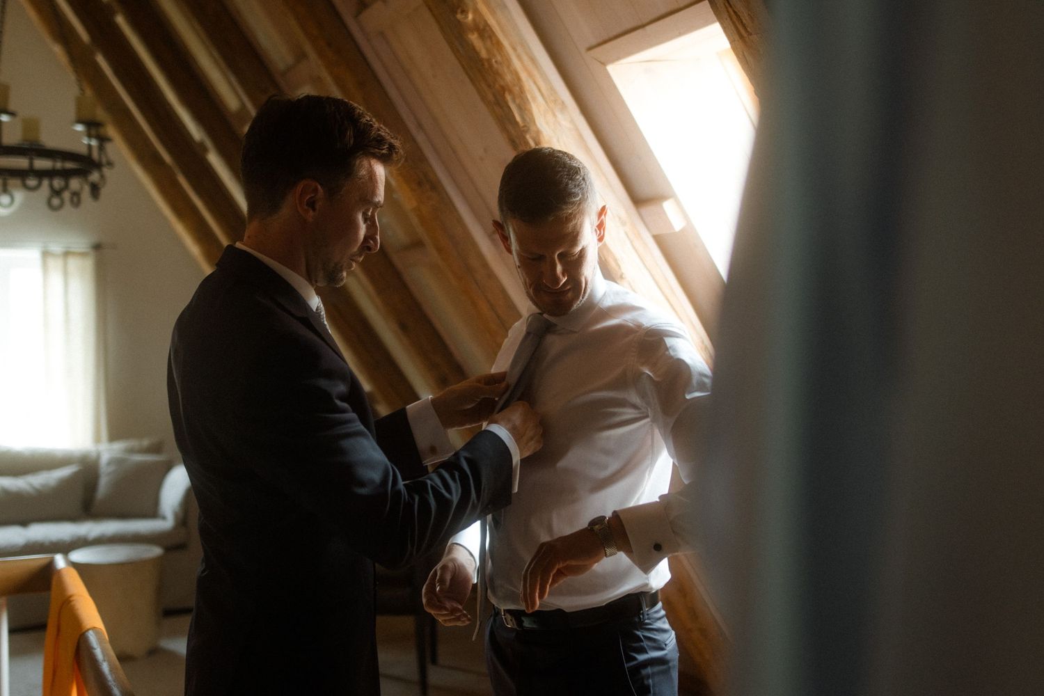 Two people adjusting formal attire in a sunlit attic space.