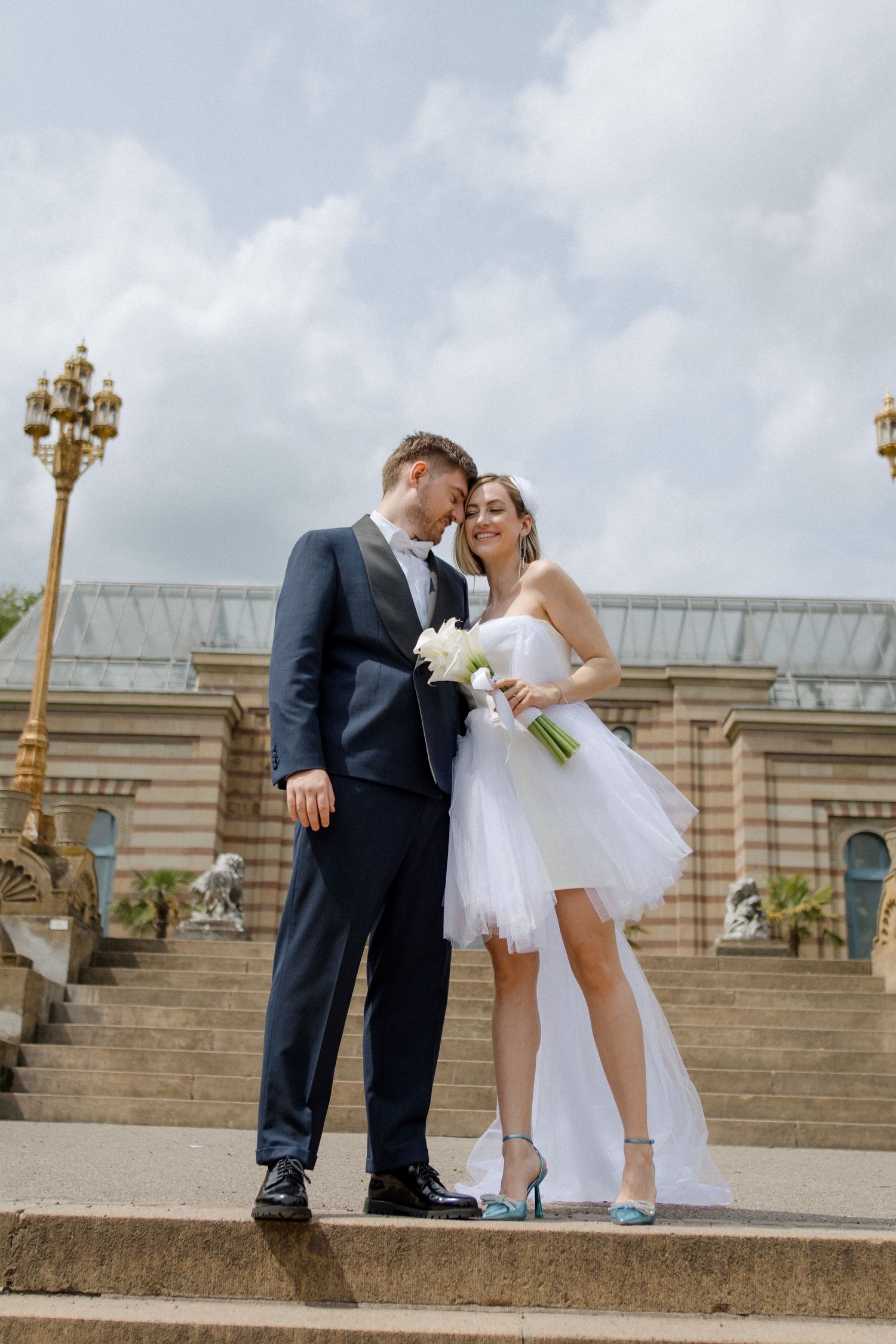 Romantic wedding couple share an intimate moment on stone steps of a classic building with ornate gold lampposts.