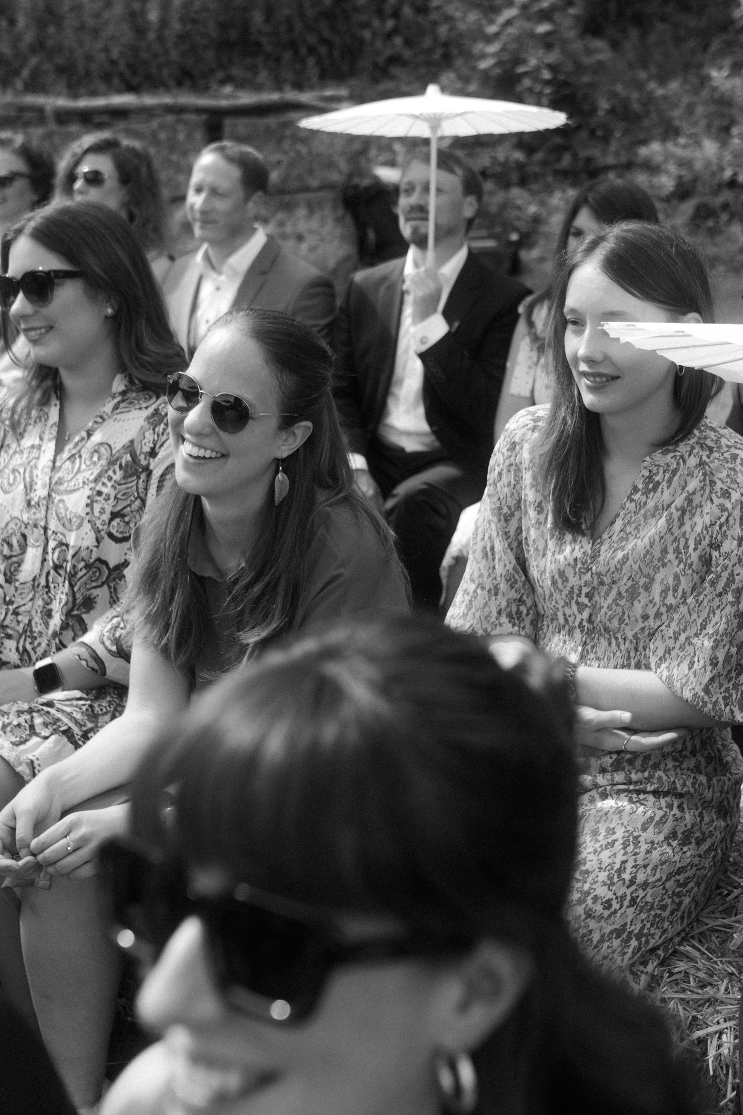 Wedding guests in formal attire sitting outdoors with parasols and sunglasses.