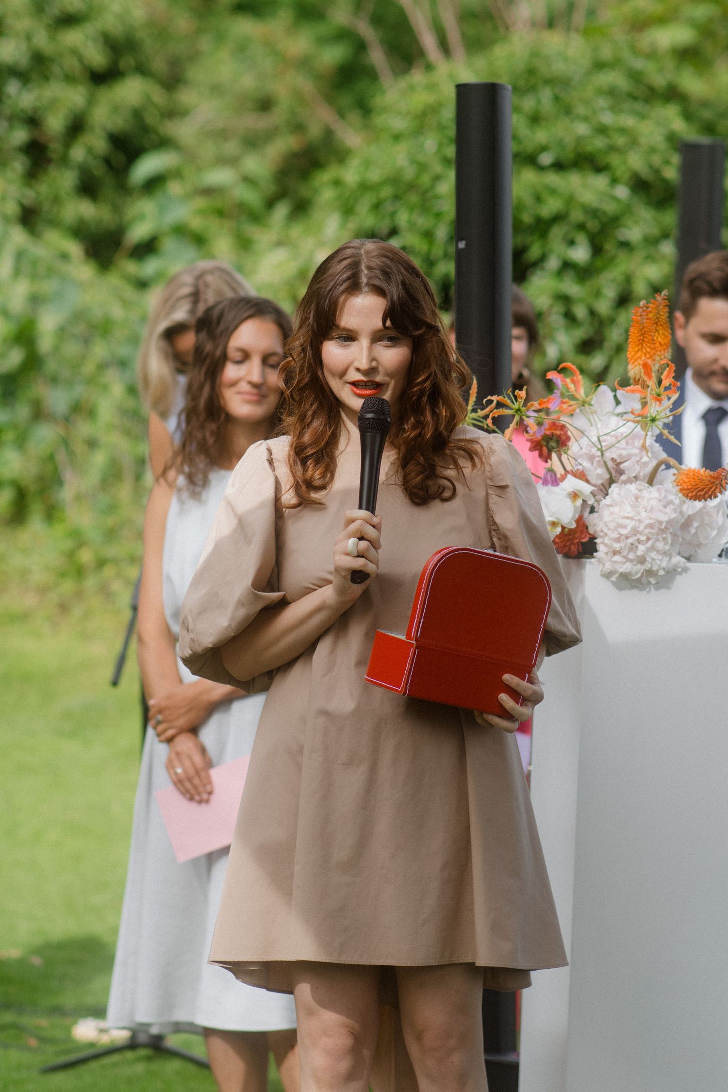 Woman in a beige dress gives a speech at an outdoor wedding reception while holding a red book.