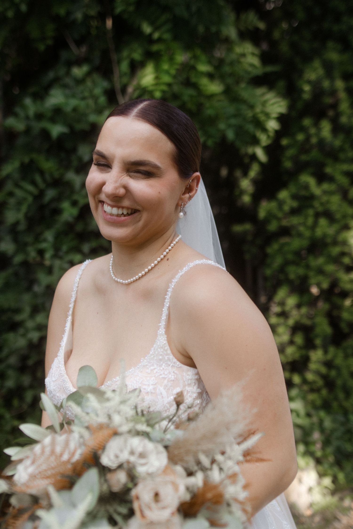 A radiant bride wearing a lace wedding dress and pearl necklace holds a romantic bouquet of roses and greenery.