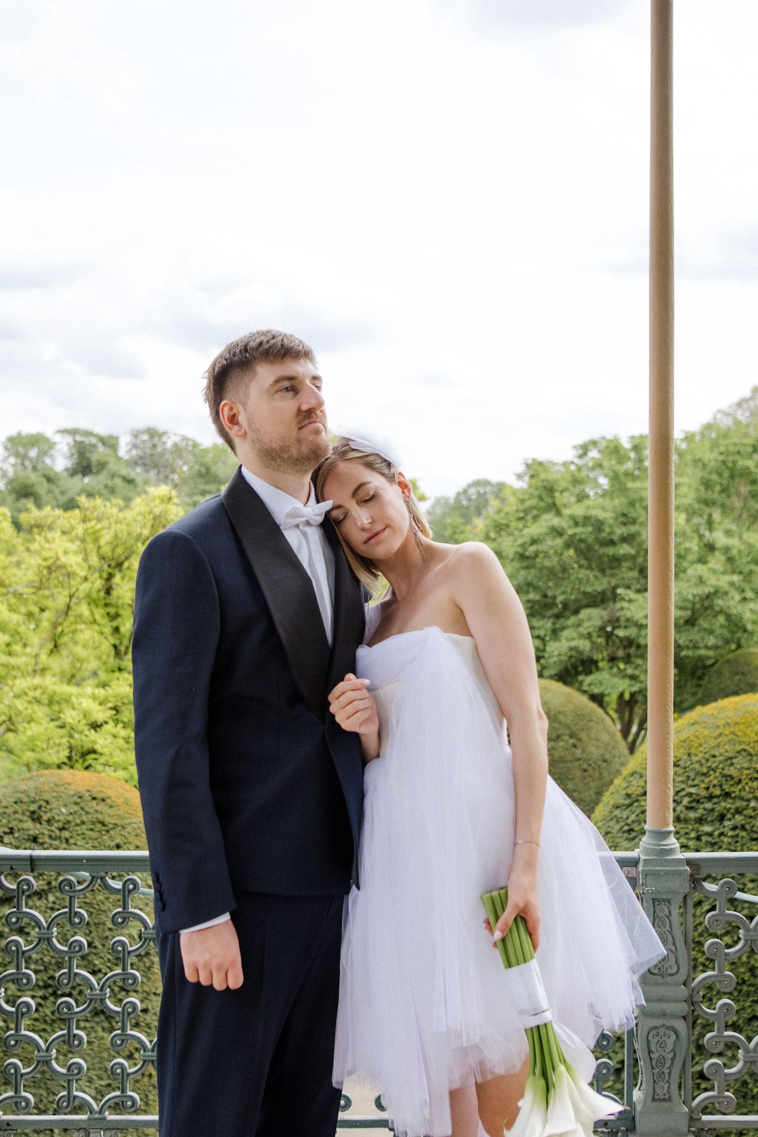 Wedding couple in formal attire standing on a balcony surrounded by greenery.