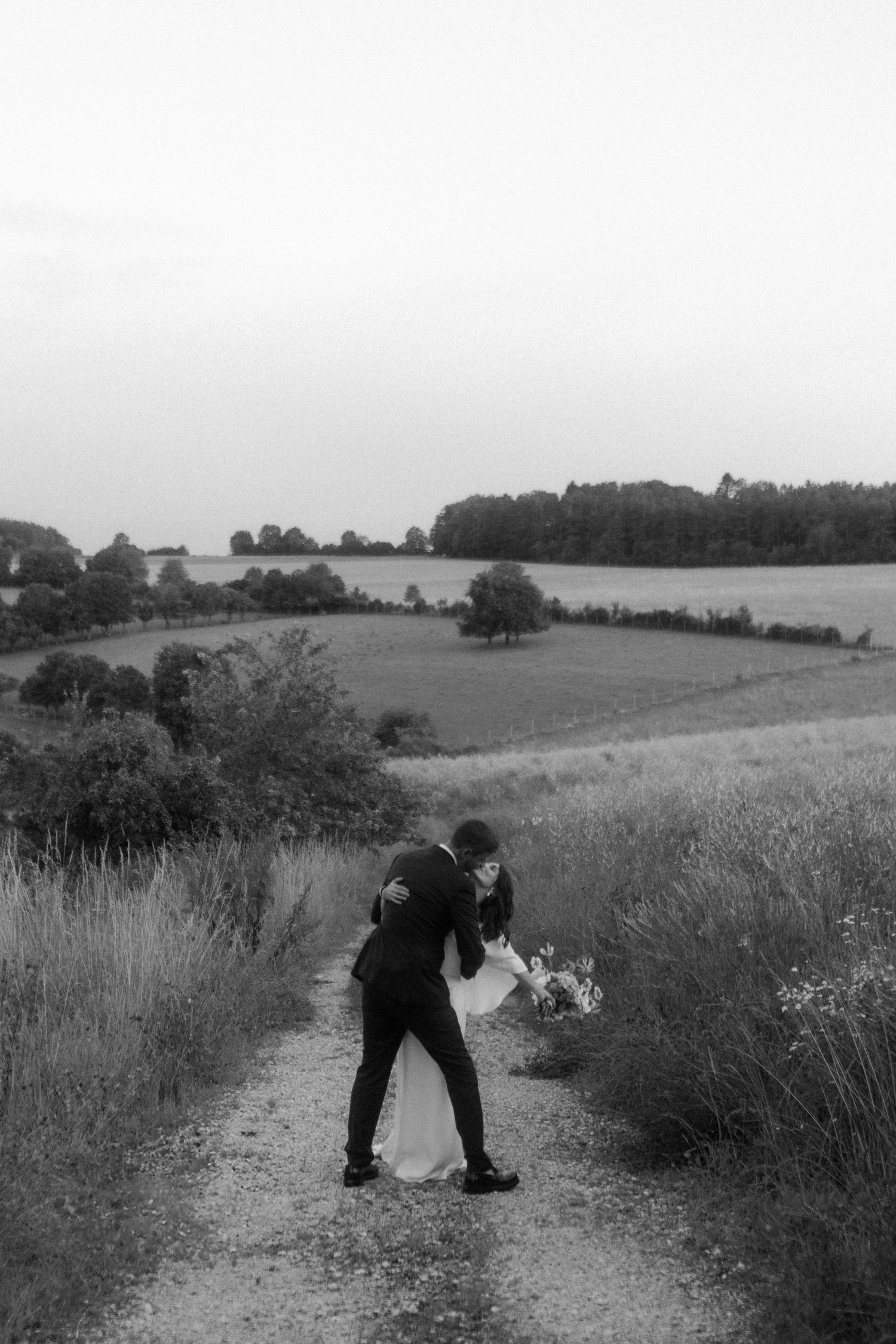 A black and white wedding photo capturing a romantic dip on a hilltop path.