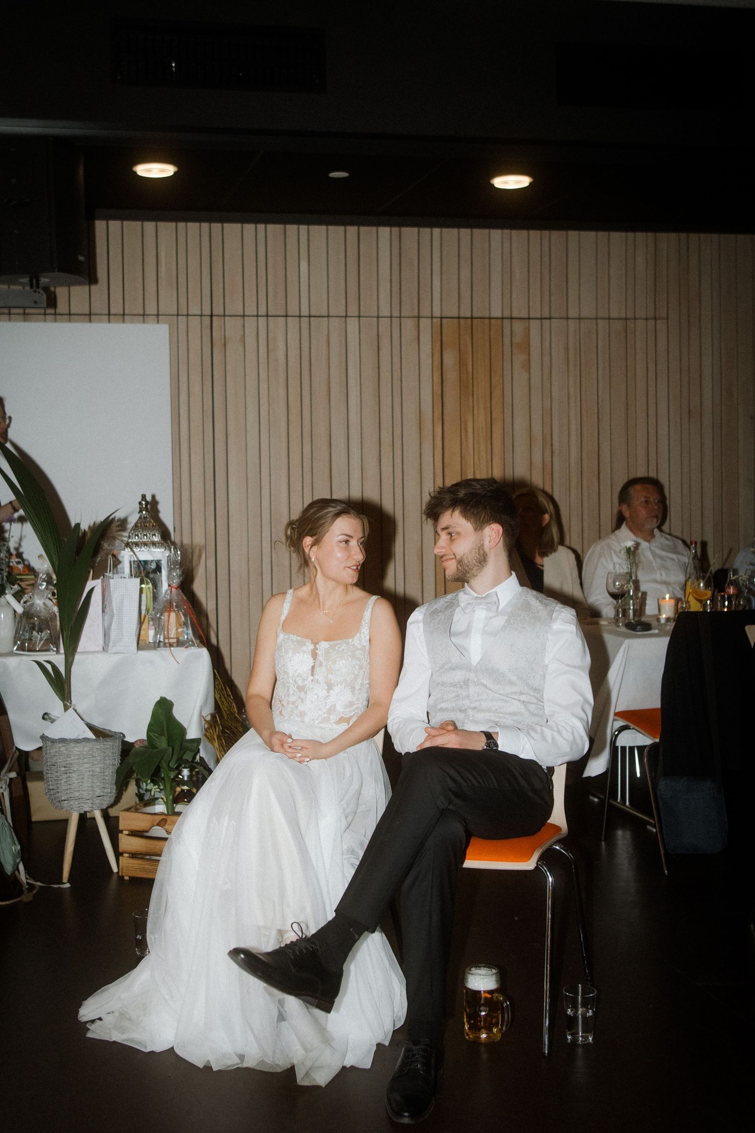 A couple sits together at their wedding reception in front of wood paneled walls.