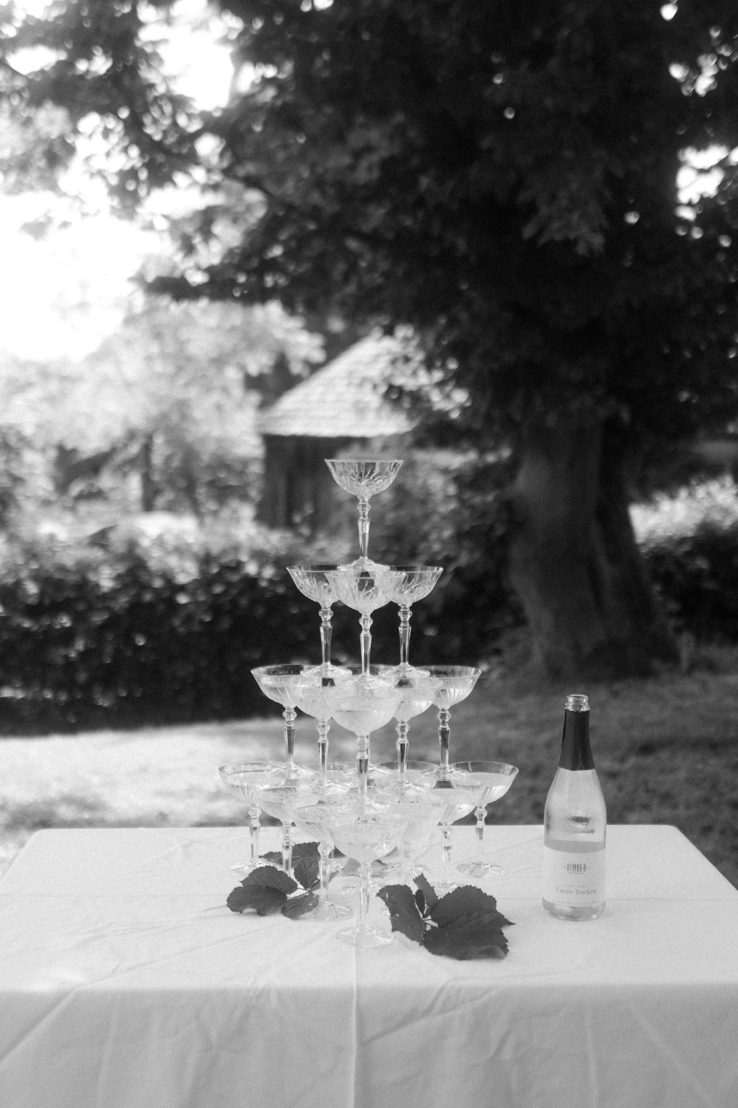 A champagne tower display with stacked crystal glasses on a white tablecloth outdoors near trees.