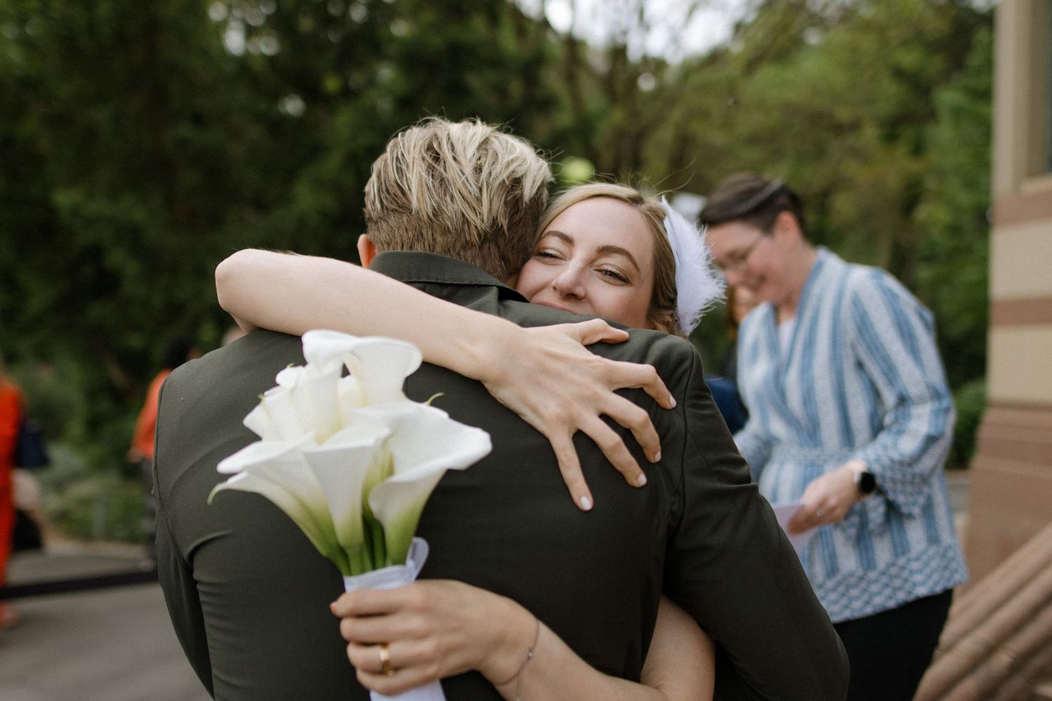 A warm embrace shared between two people holding white calla lilies at an outdoor gathering.