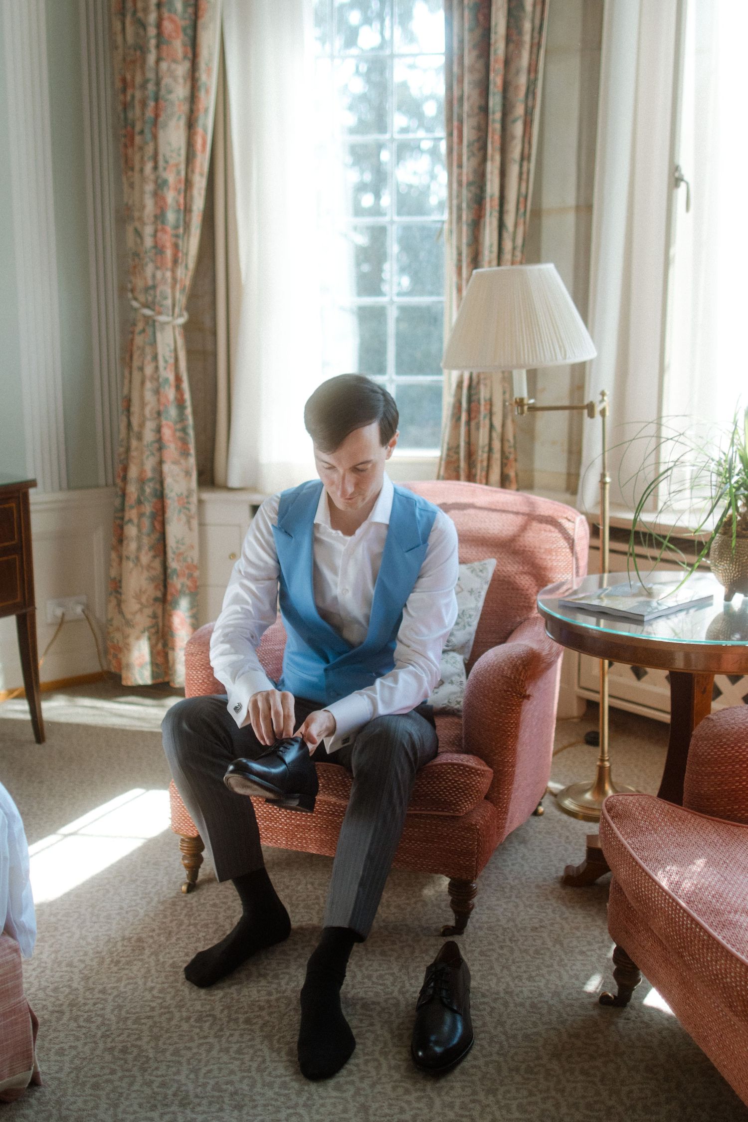 Person in blue vest sitting on pink couch near sunlit windows in elegant room.