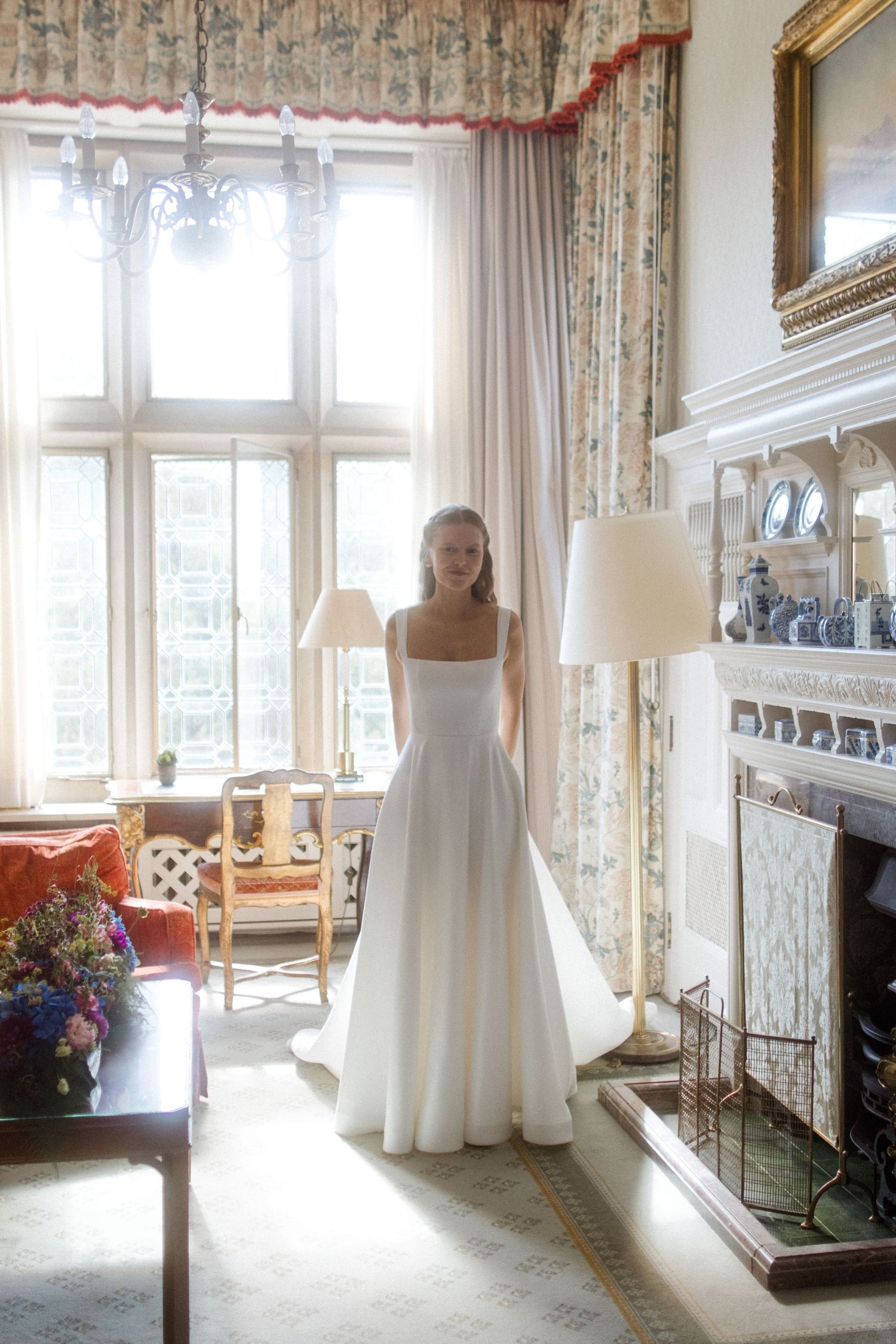 White wedding dress displayed in sunlit room with floral curtains and elegant furniture.