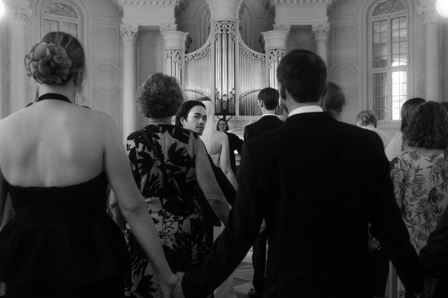 Elegant wedding guests gather around a grand organ in a classical white interior church setting.
