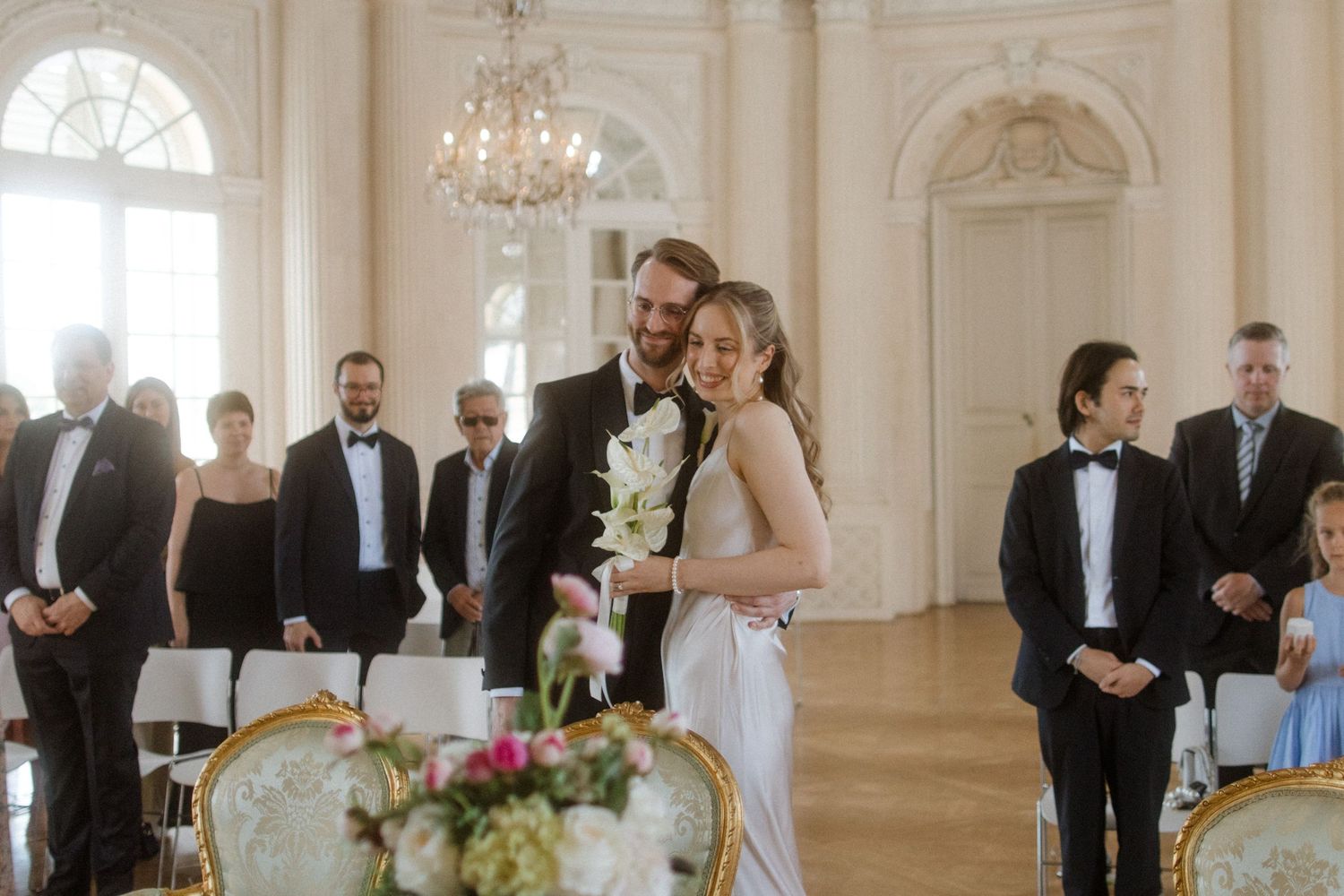 An intimate wedding ceremony in a luxurious white baroque hall with chandeliers.