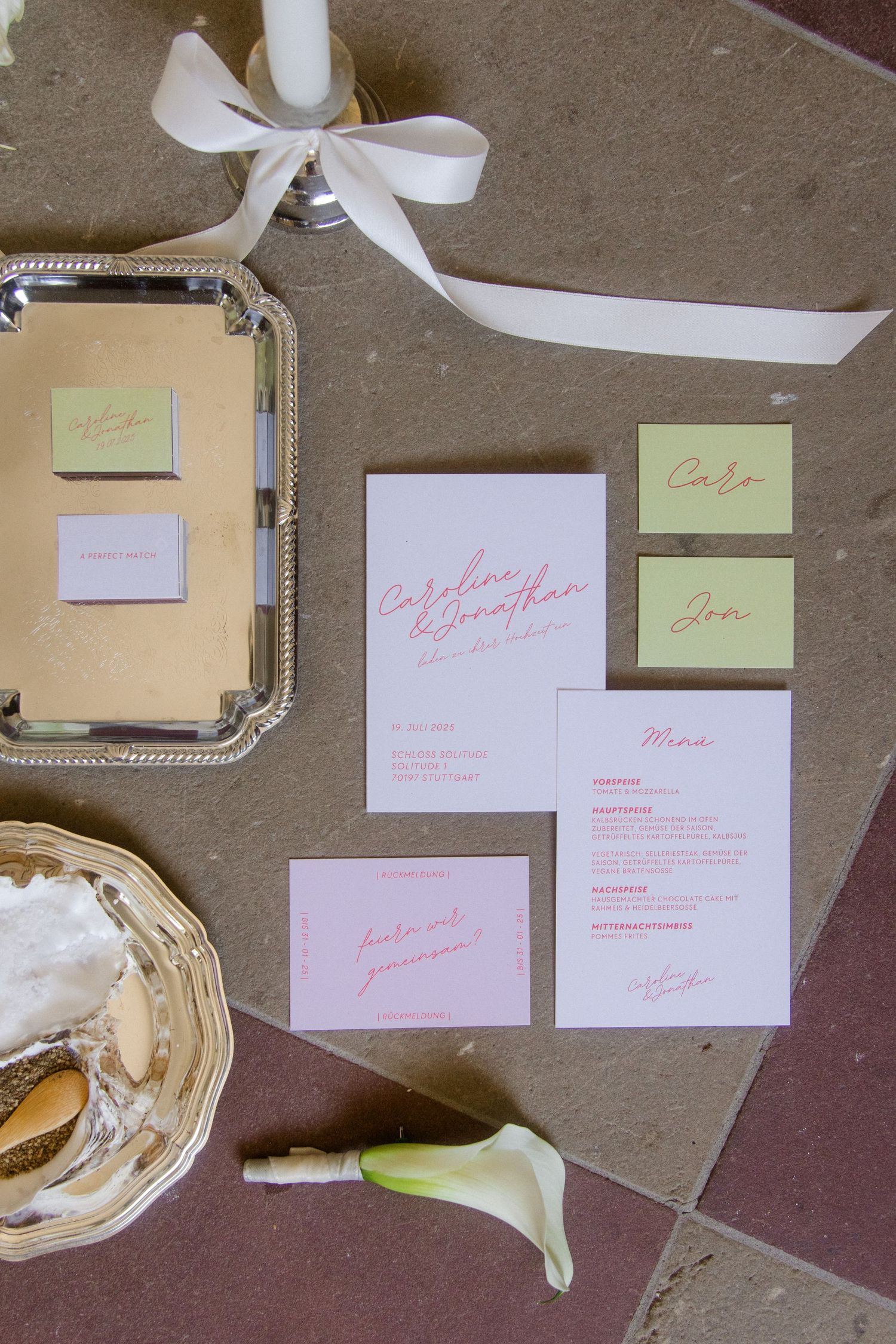 Wedding invitation suite with pink lettering and metallic accents laid out on brown surface.