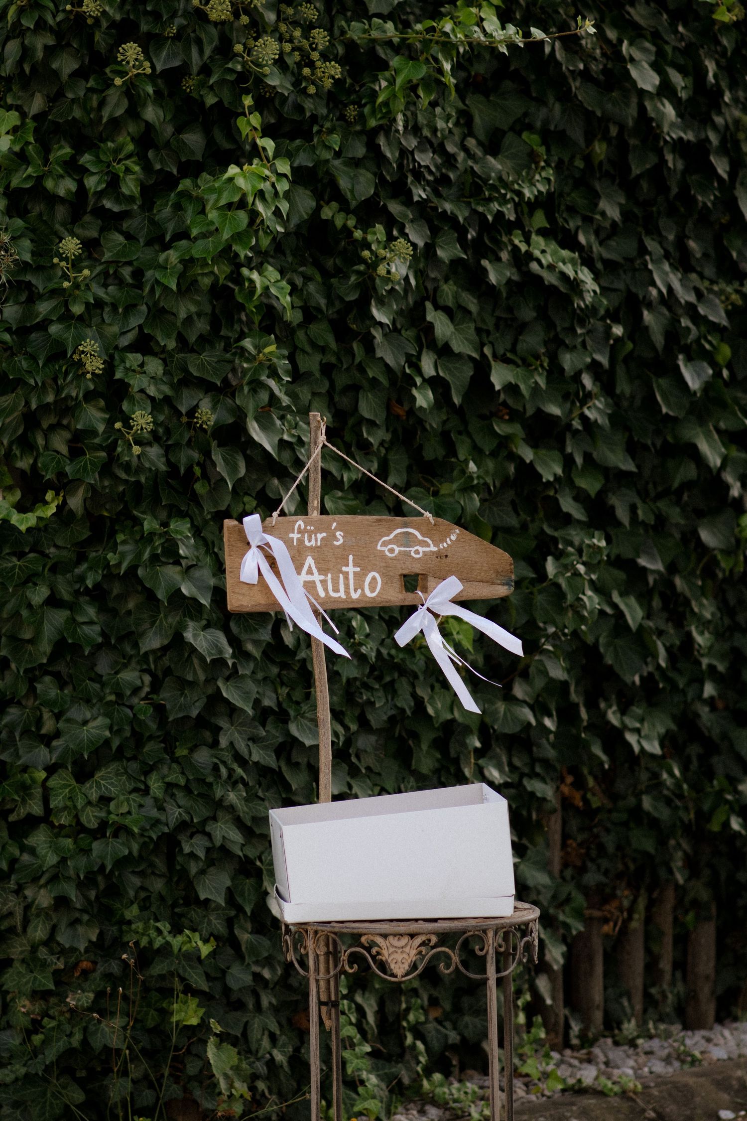 Rustic wooden wedding signs with white ribbons displayed against a leafy green hedge background.