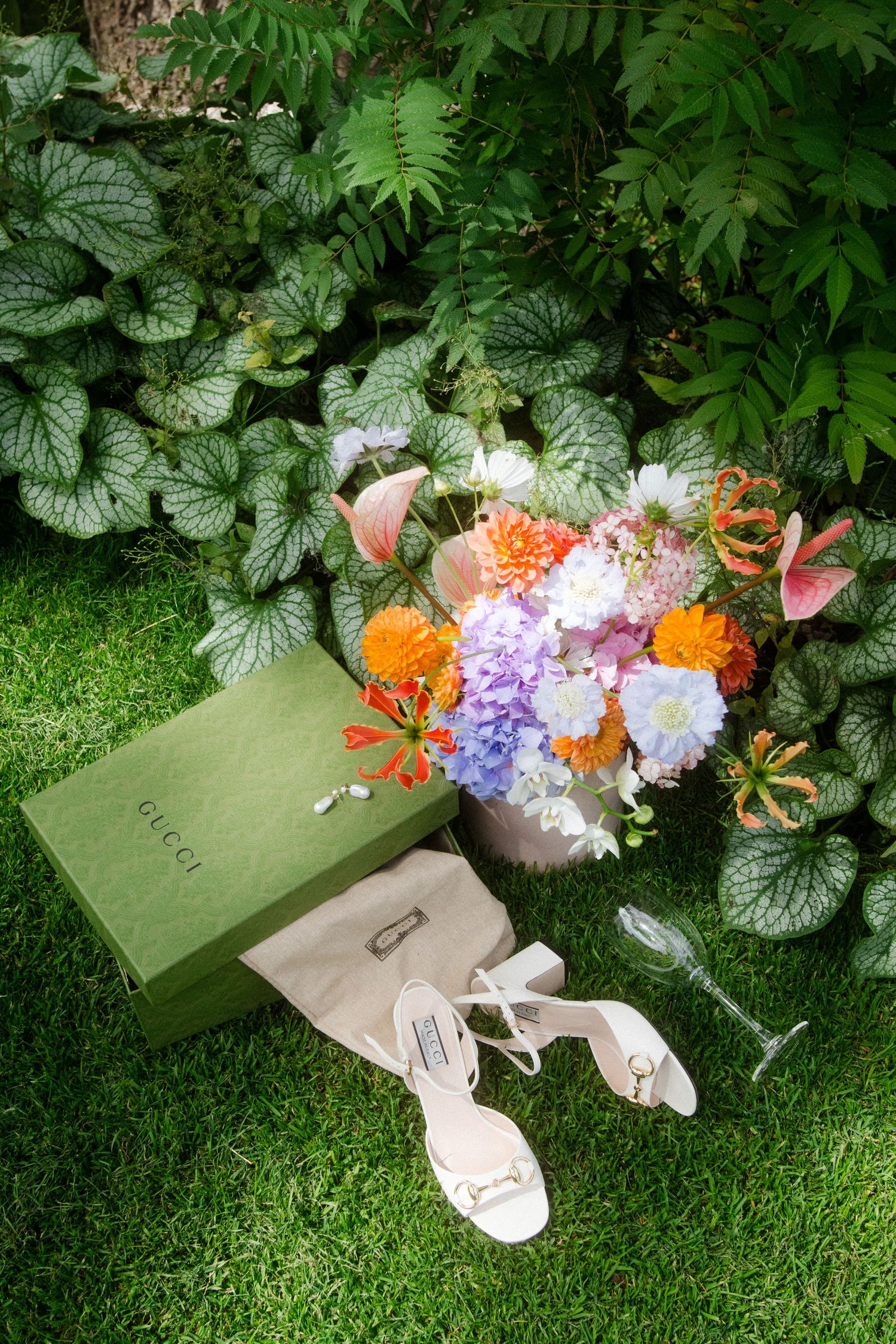 Wedding shoes and floral arrangements displayed on green grass with lush foliage background.