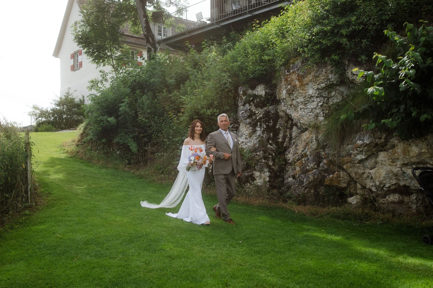 A couple poses for a wedding photo against a stone wall and lush greenery.