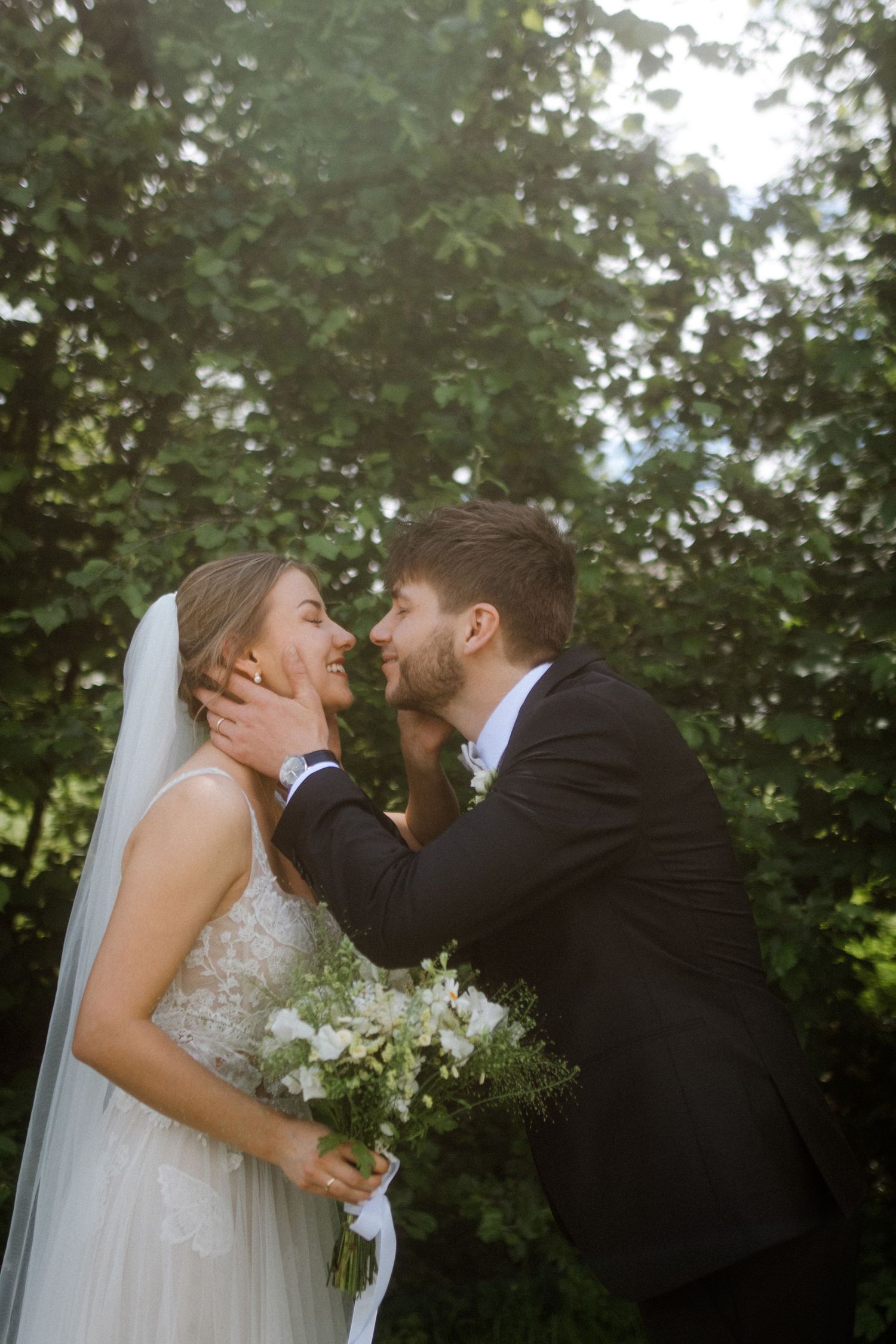 Newlywed couple shares a romantic kiss outdoors surrounded by greenery.