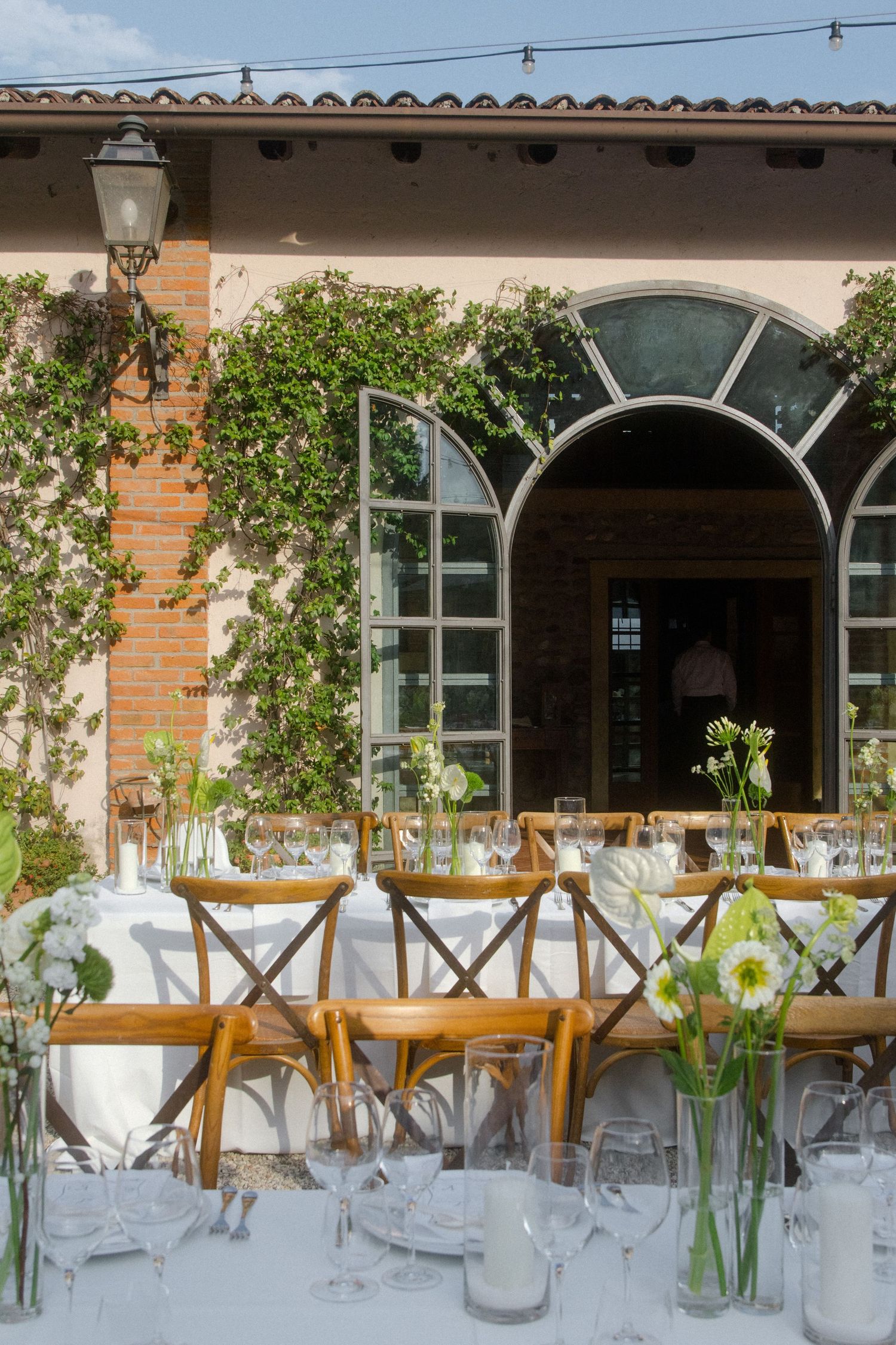 Elegant outdoor dining setup with white tablecloths and glass vases under arched windows and climbing vines.