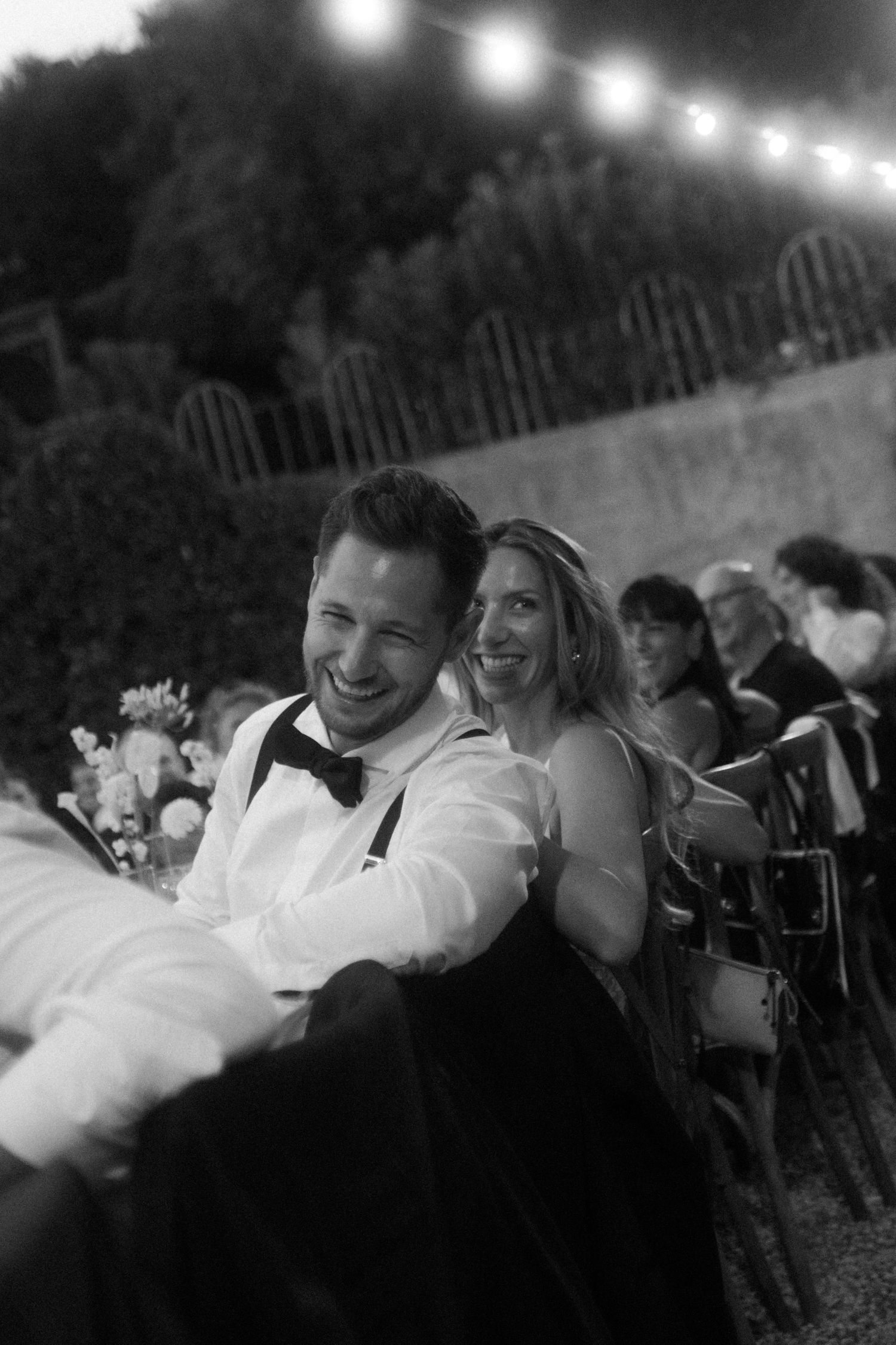 Black and white candid photo of guests enjoying a wedding reception under evening lights.
