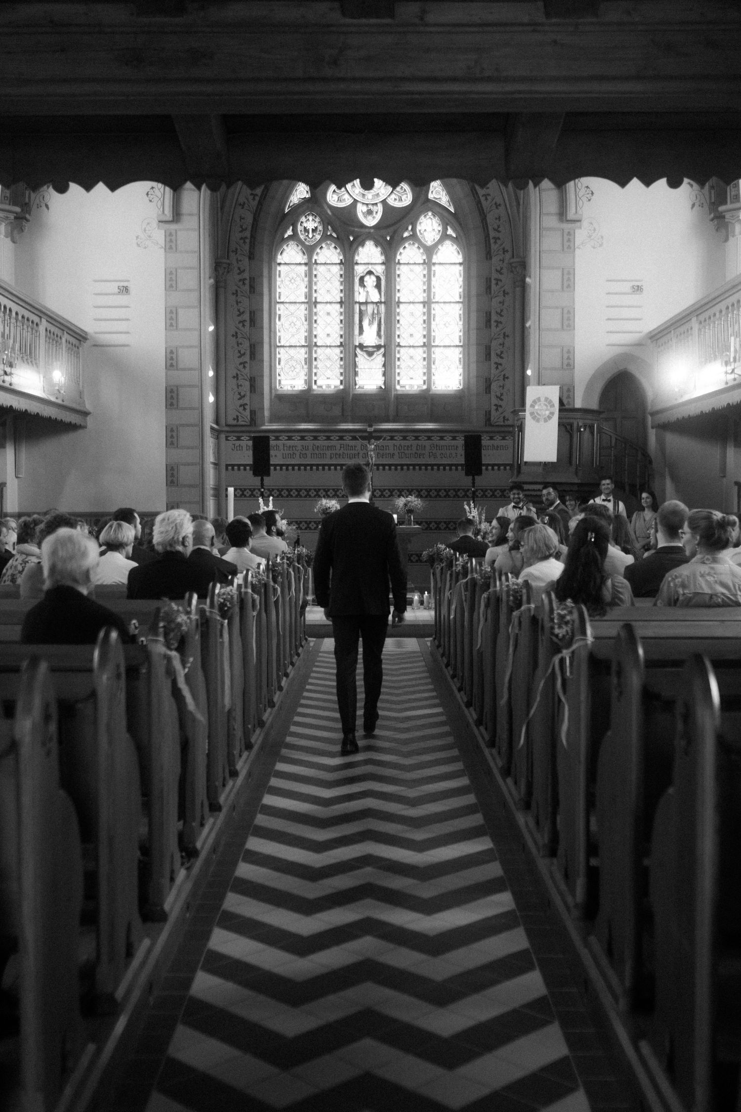 Black and white photo of church aisle with chevron pattern leading to stained glass window.