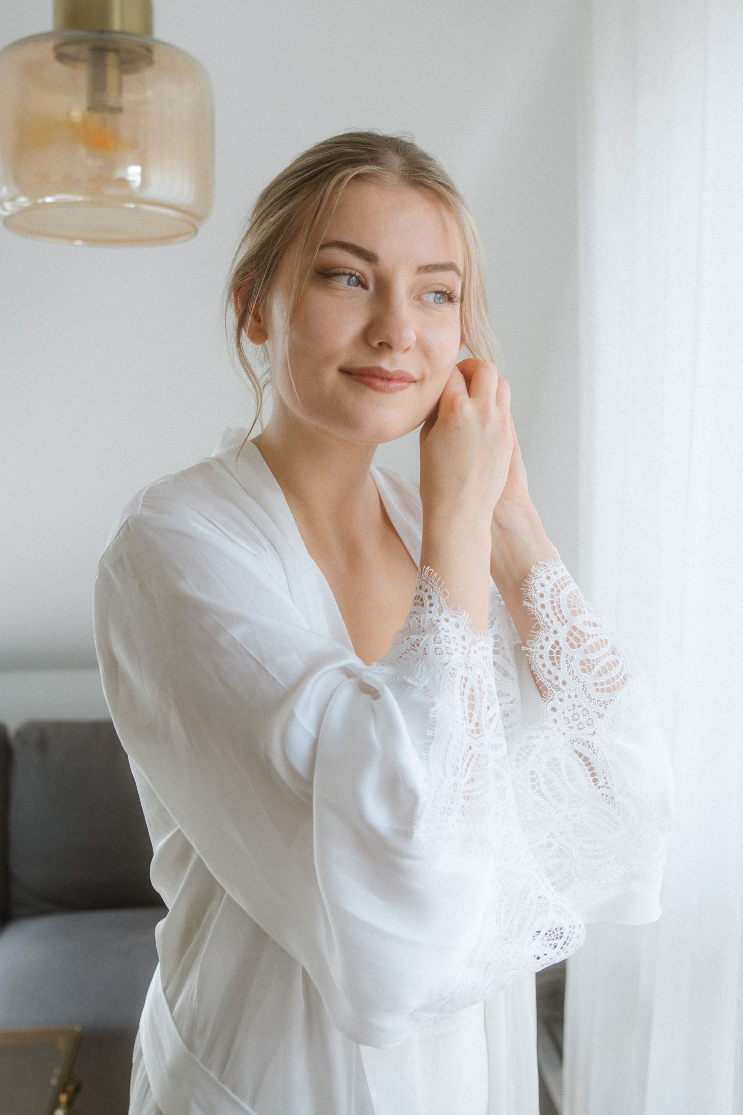 Woman in white lace robe standing by window in soft morning light.