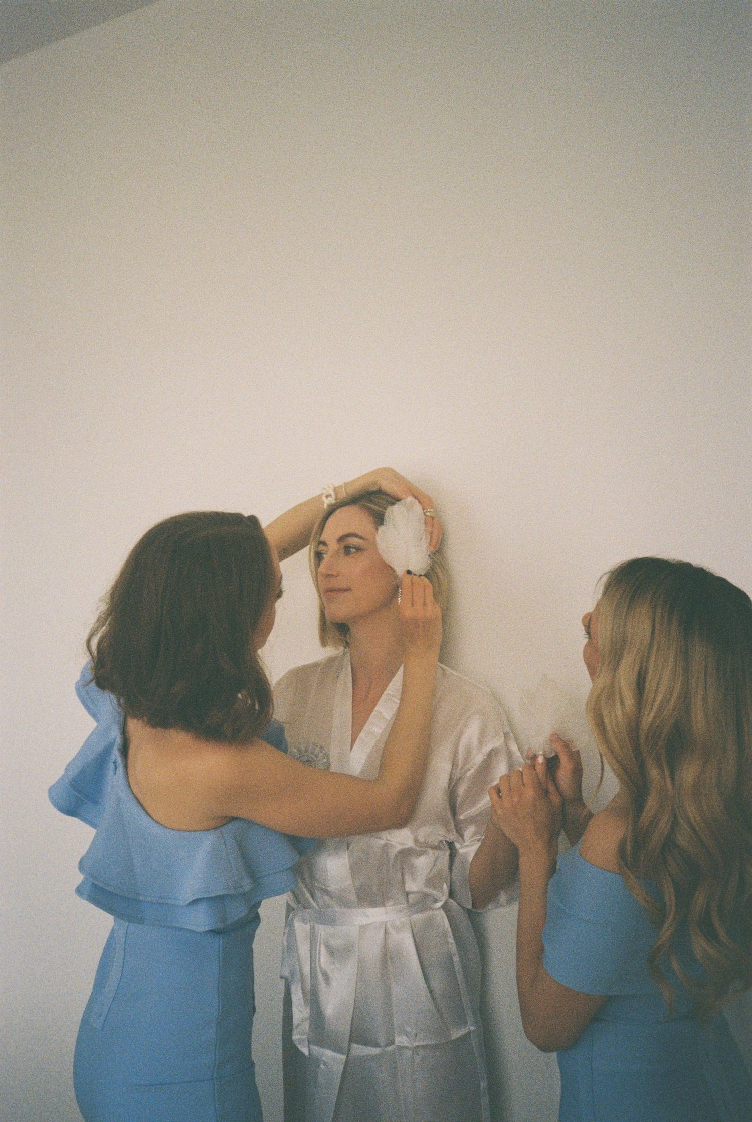 Getting ready scene with bridesmaids in blue dresses helping the bride prepare.