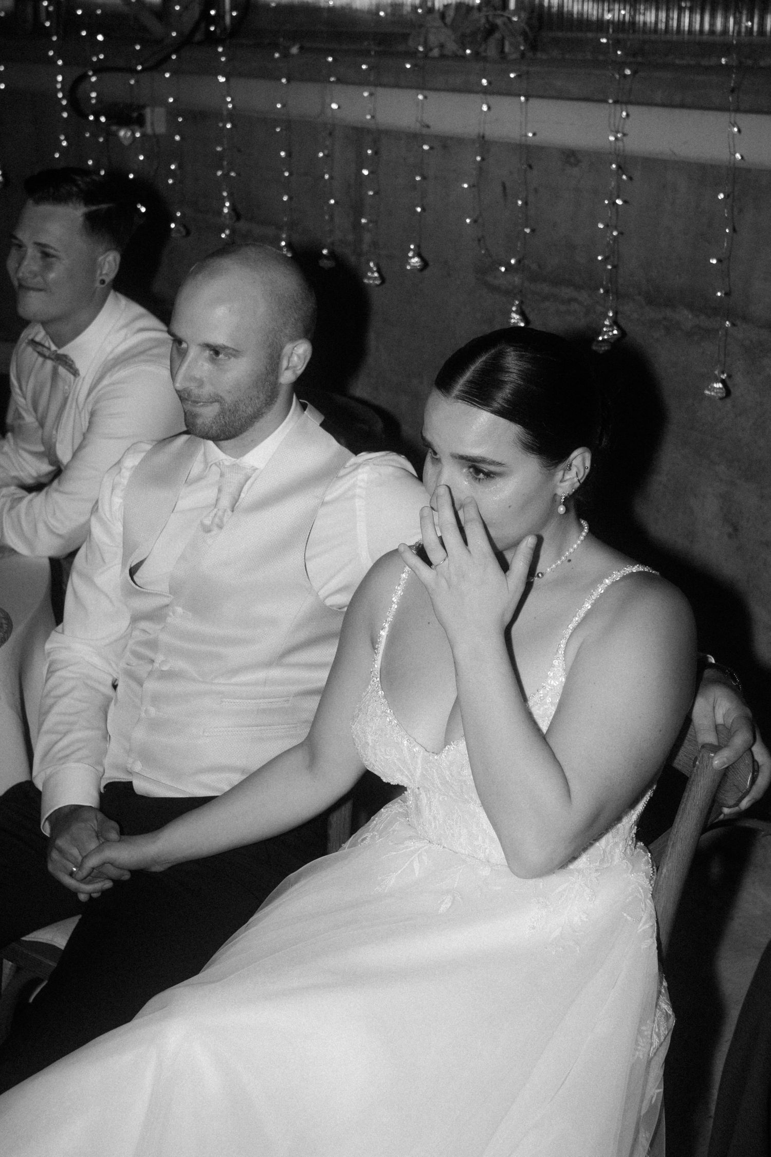 Wedding guests seated at reception react emotionally to a speech under twinkling string lights.
