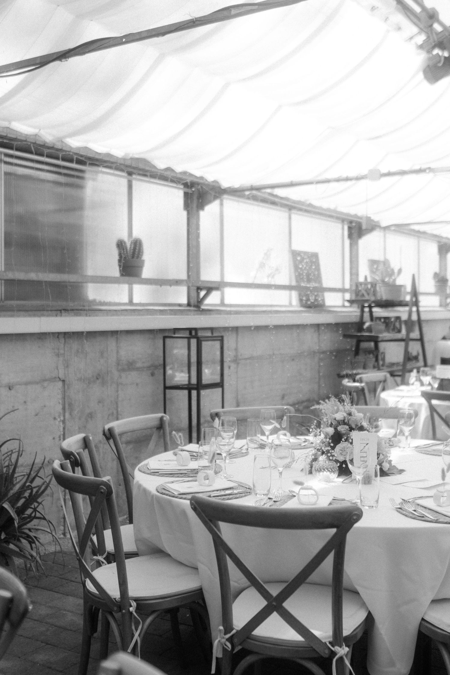 A black and white interior of a restaurant with round tables set with white tablecloths and cross-back chairs.