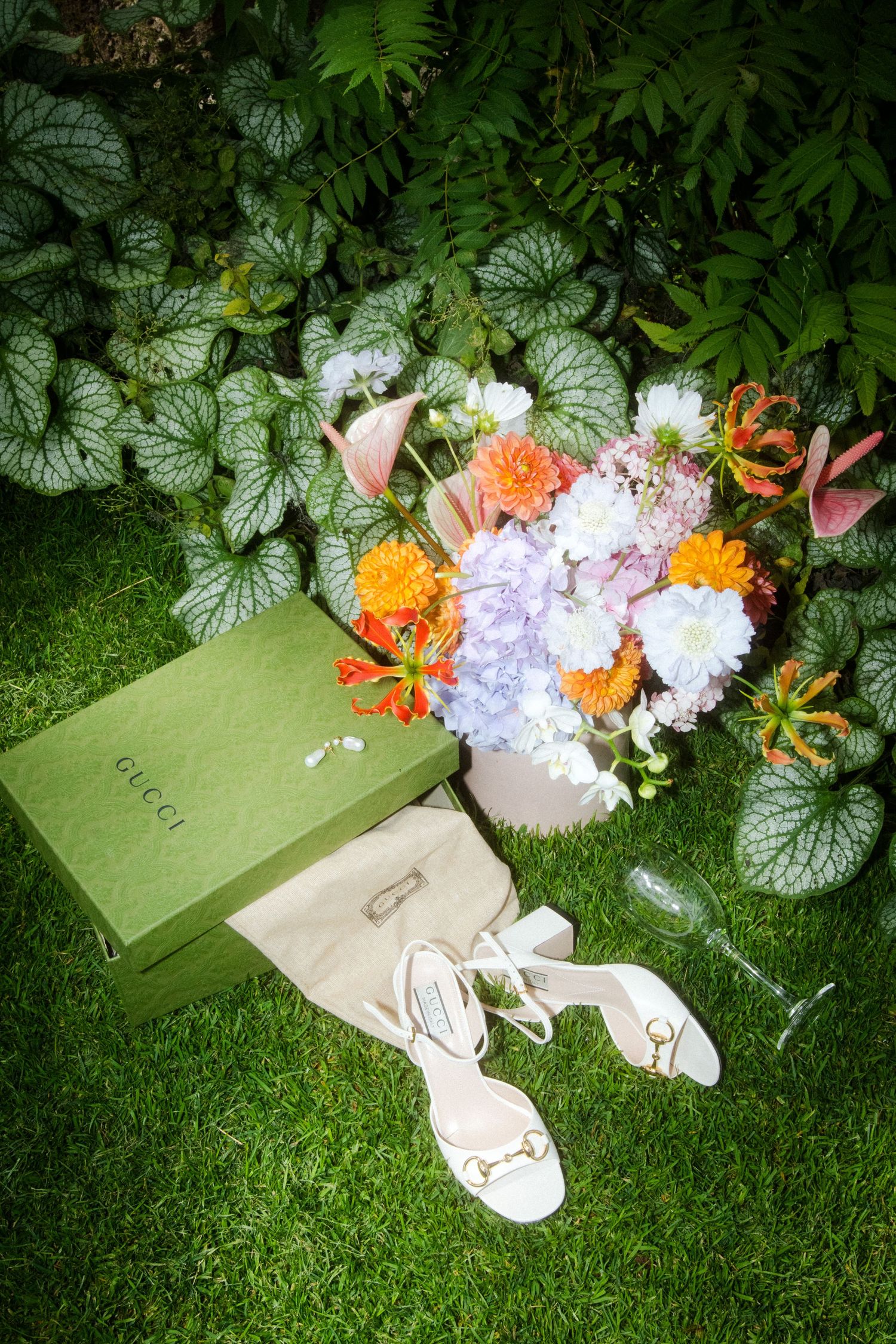 Wedding accessories including shoes and flowers arranged on grass with decorative foliage.