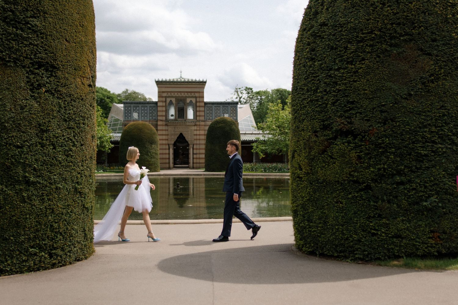 Garden scene with formal hedges framing a classical building reflection in a pond.