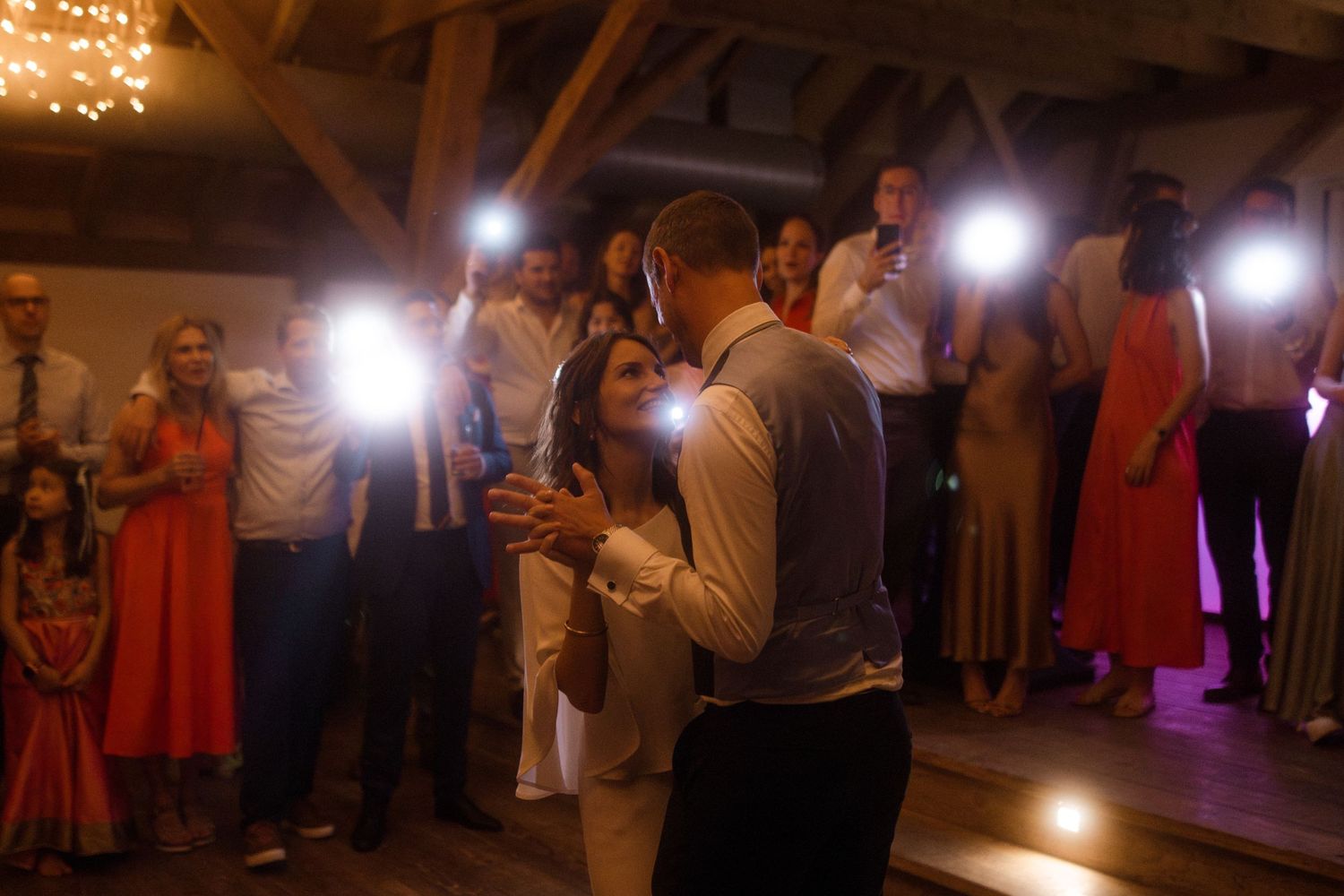 Wedding guests surround a dancing couple at an indoor reception with warm lighting.