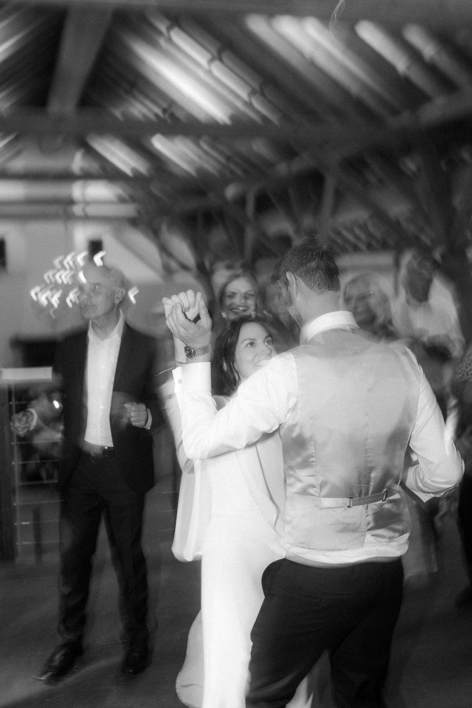 Wedding guests dancing and celebrating at a reception under decorative lighting in black and white.