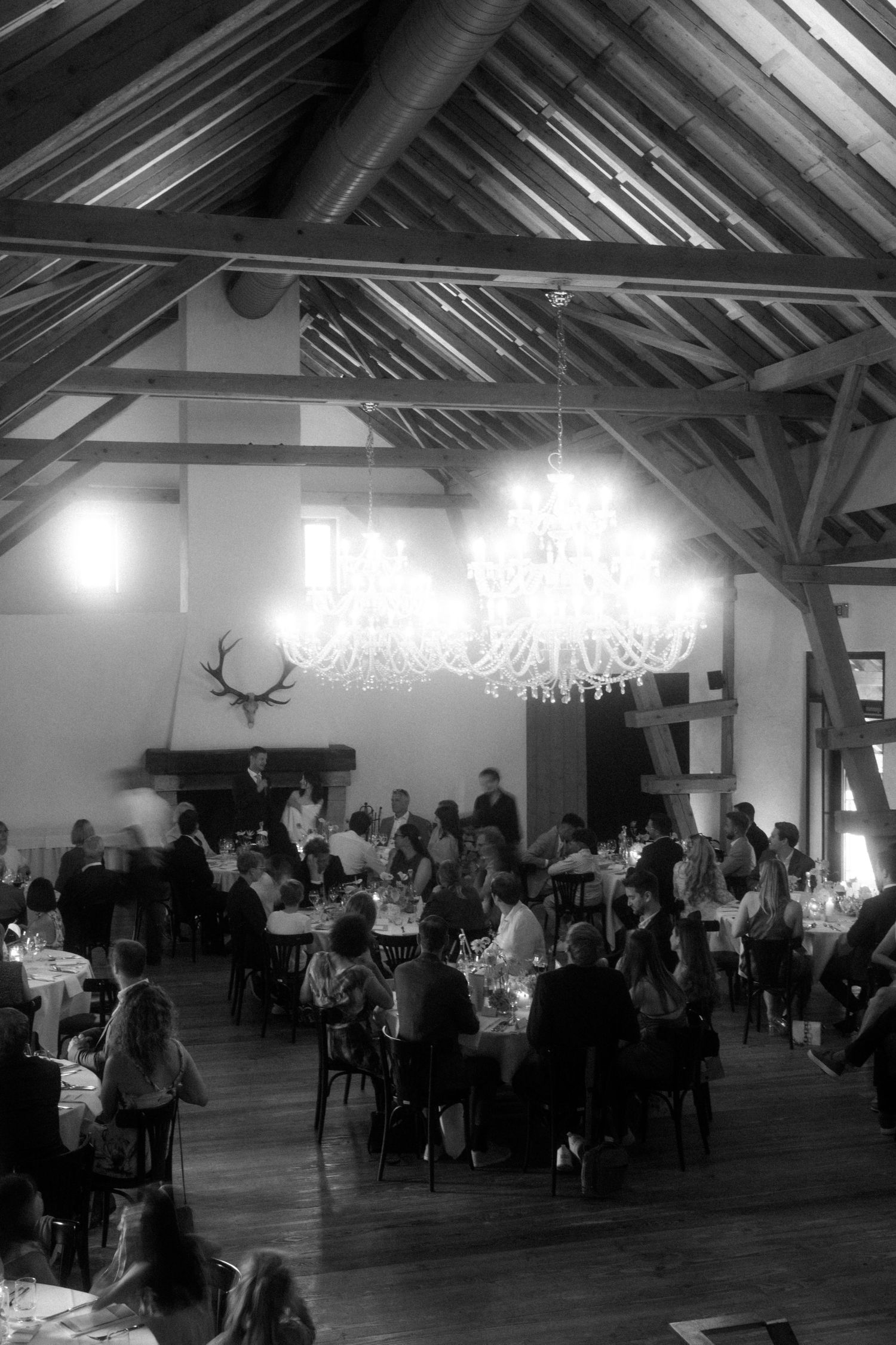 Black and white photo of wedding reception with chandelier and antler decor in rustic venue.
