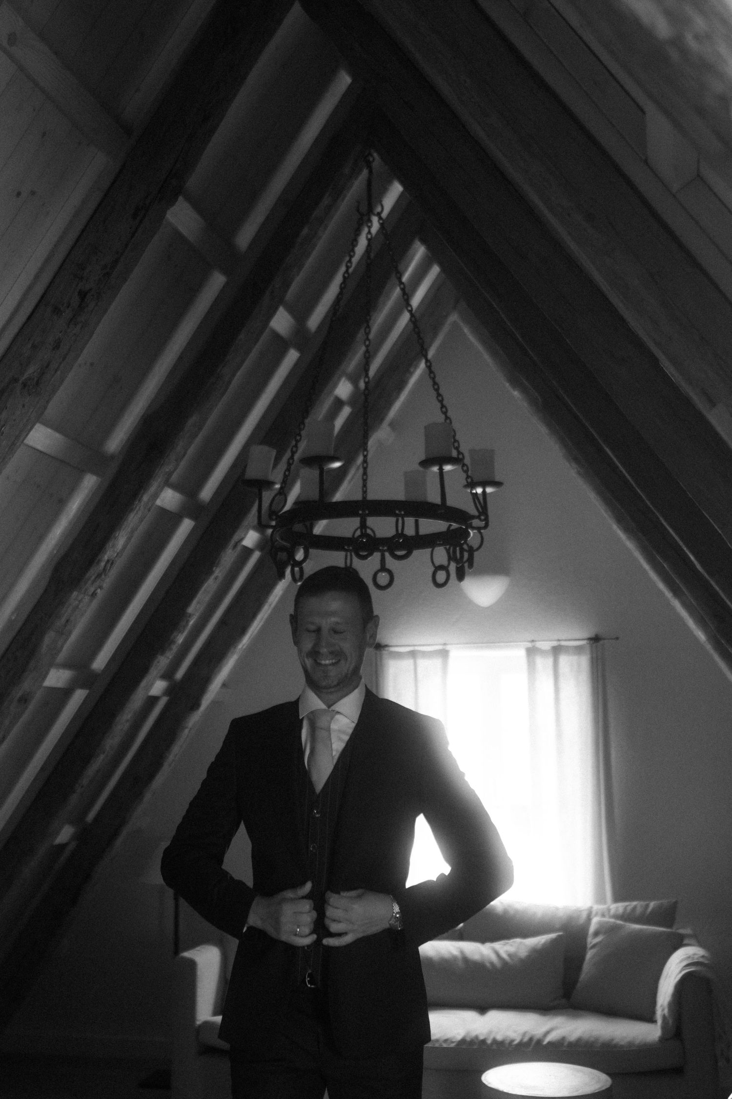 Figure in dark suit standing in attic room with chandelier and sunlight streaming through window.