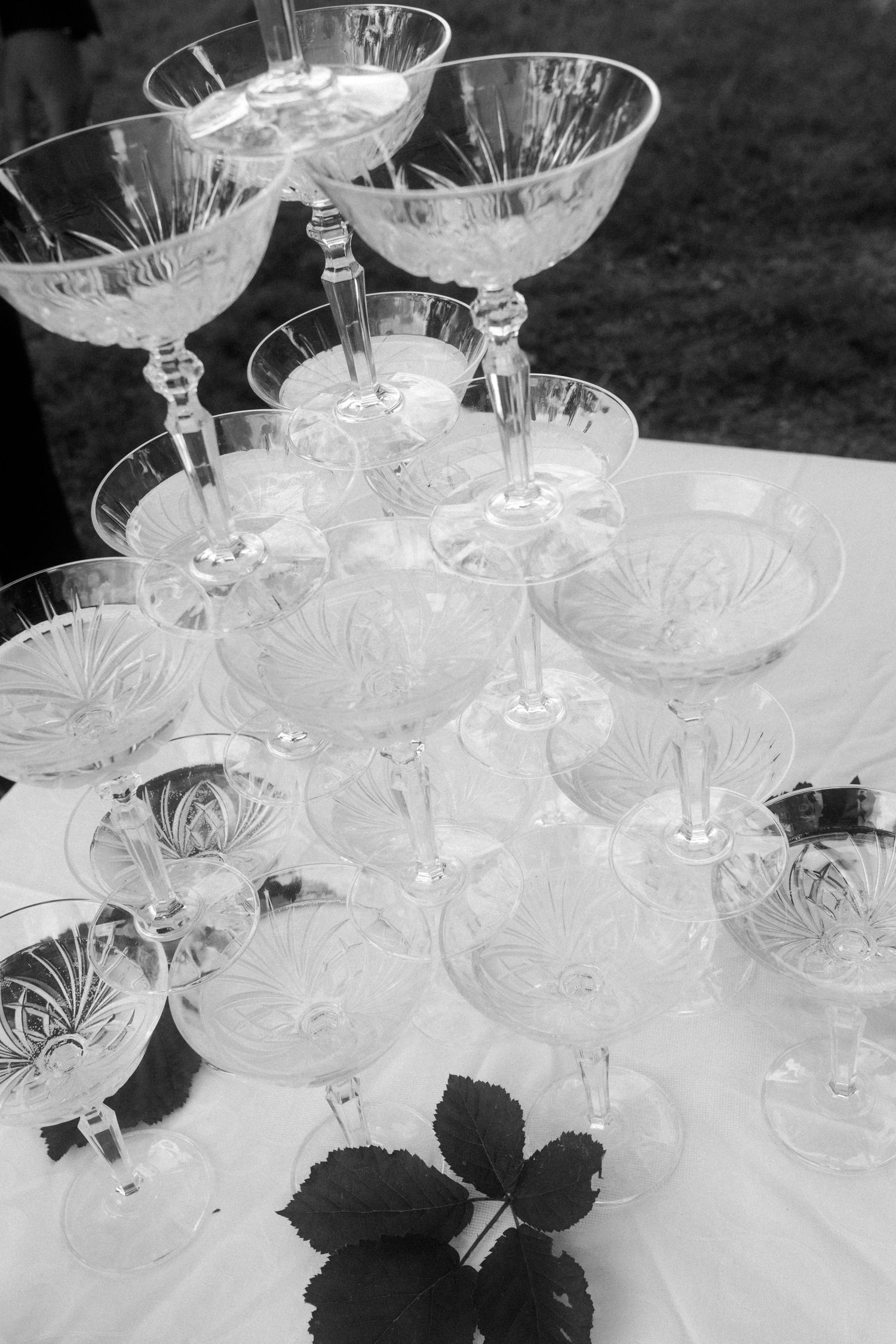 Elegant crystal coupe glasses arranged in tower formation on white linen with decorative leaves.