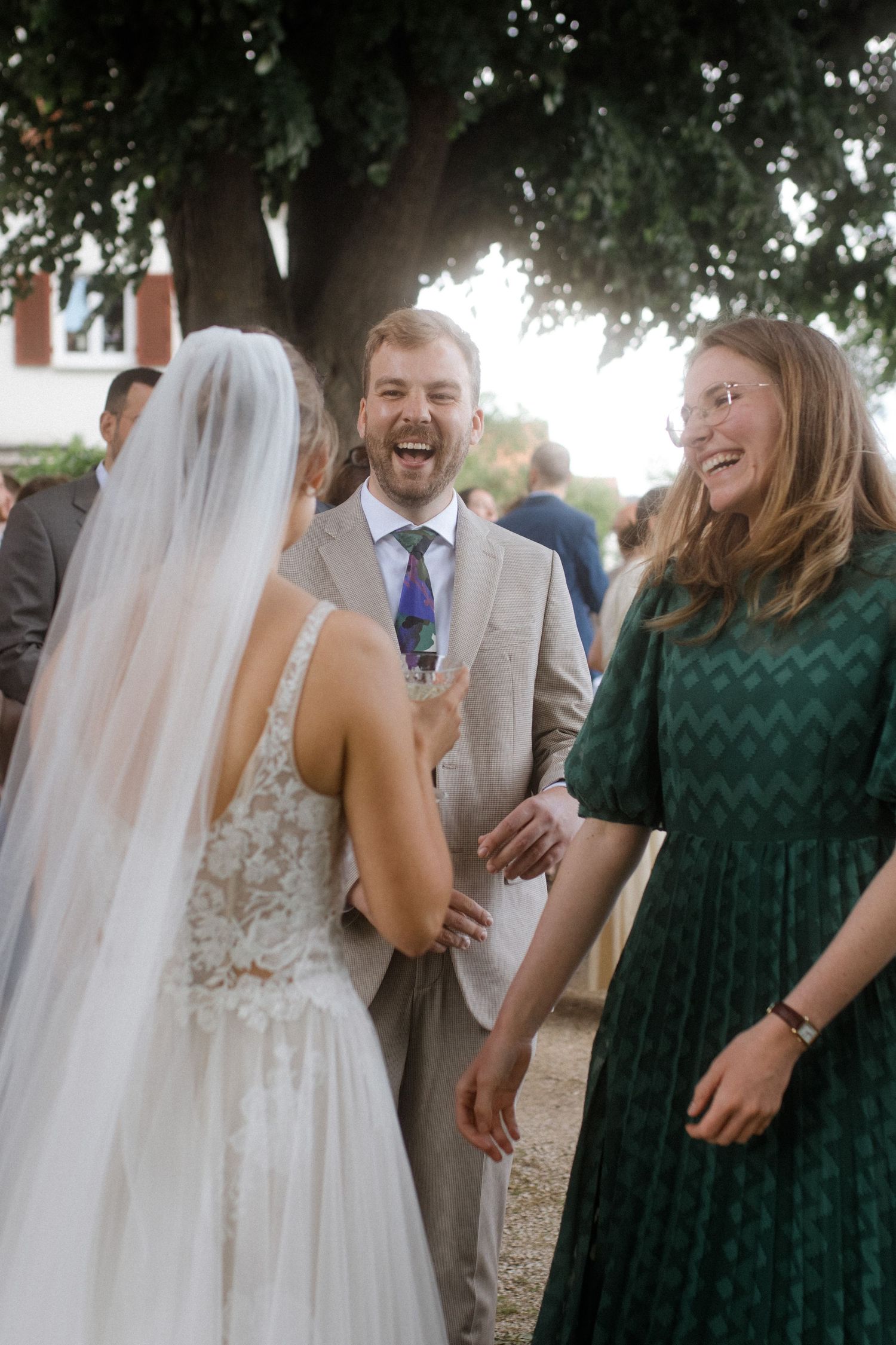 Bride in white dress shares laughter with wedding guests under tree.