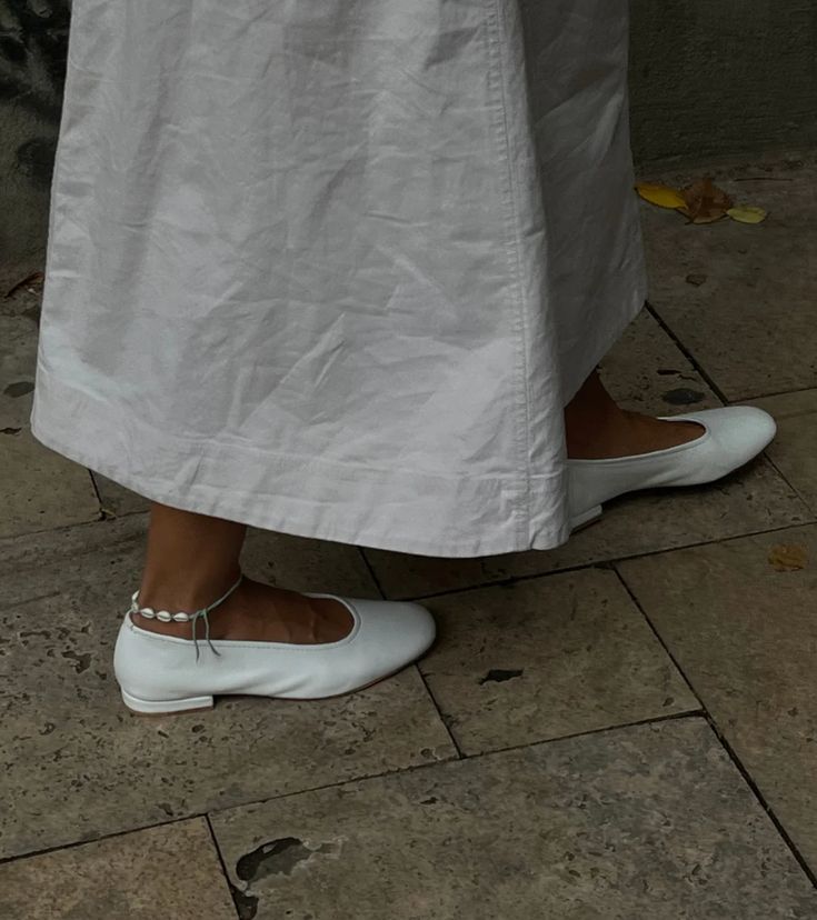 Classic white ballet flats peeking out from under a long white dress.