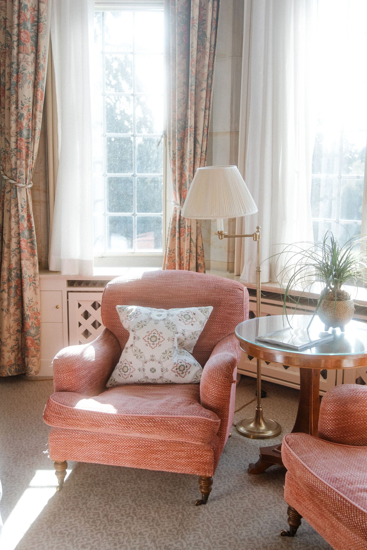 Elegant coral-colored armchairs arranged in a bright sunlit room with floral curtains.