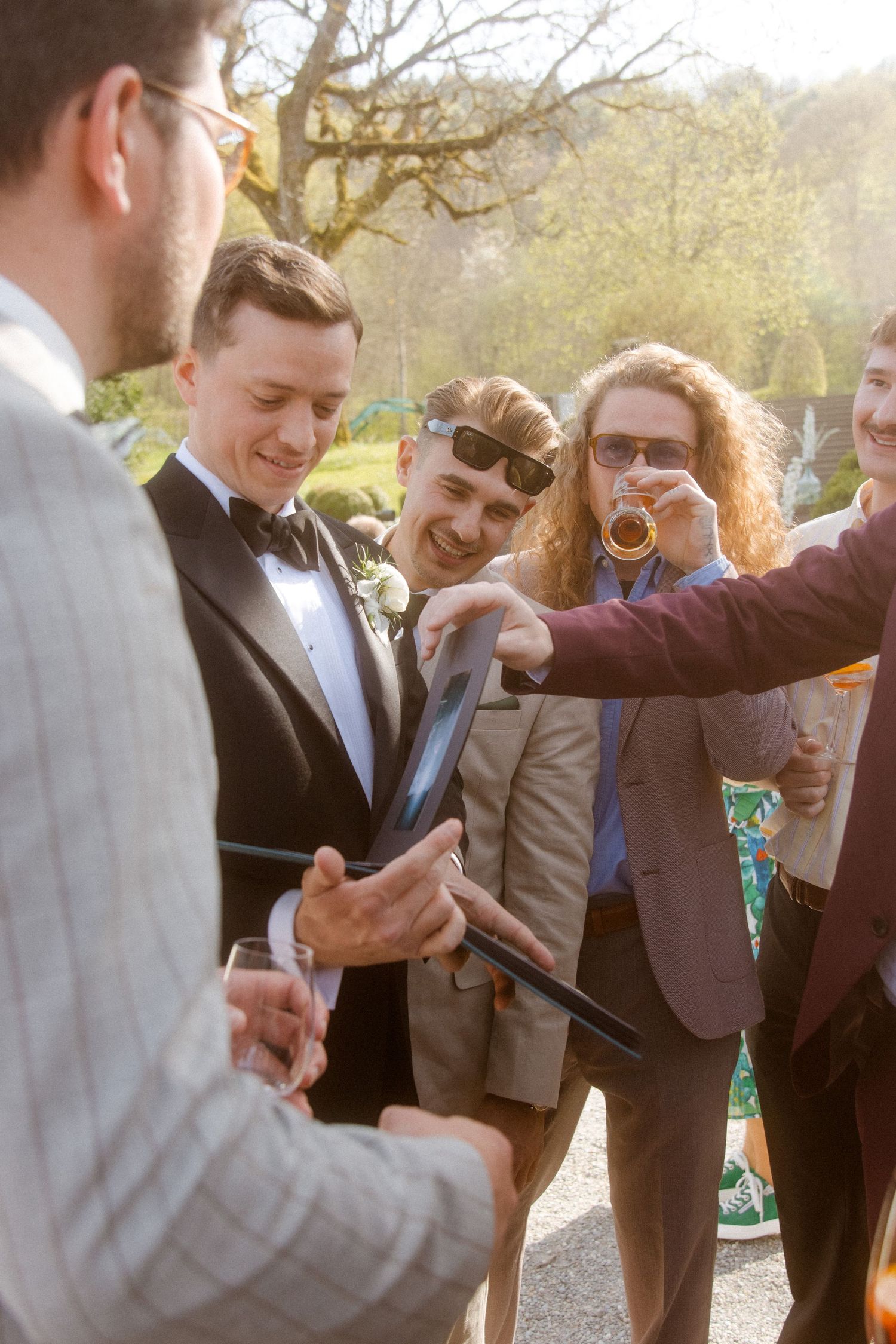 Groom in tuxedo with friends examining item at sunny outdoor wedding reception.
