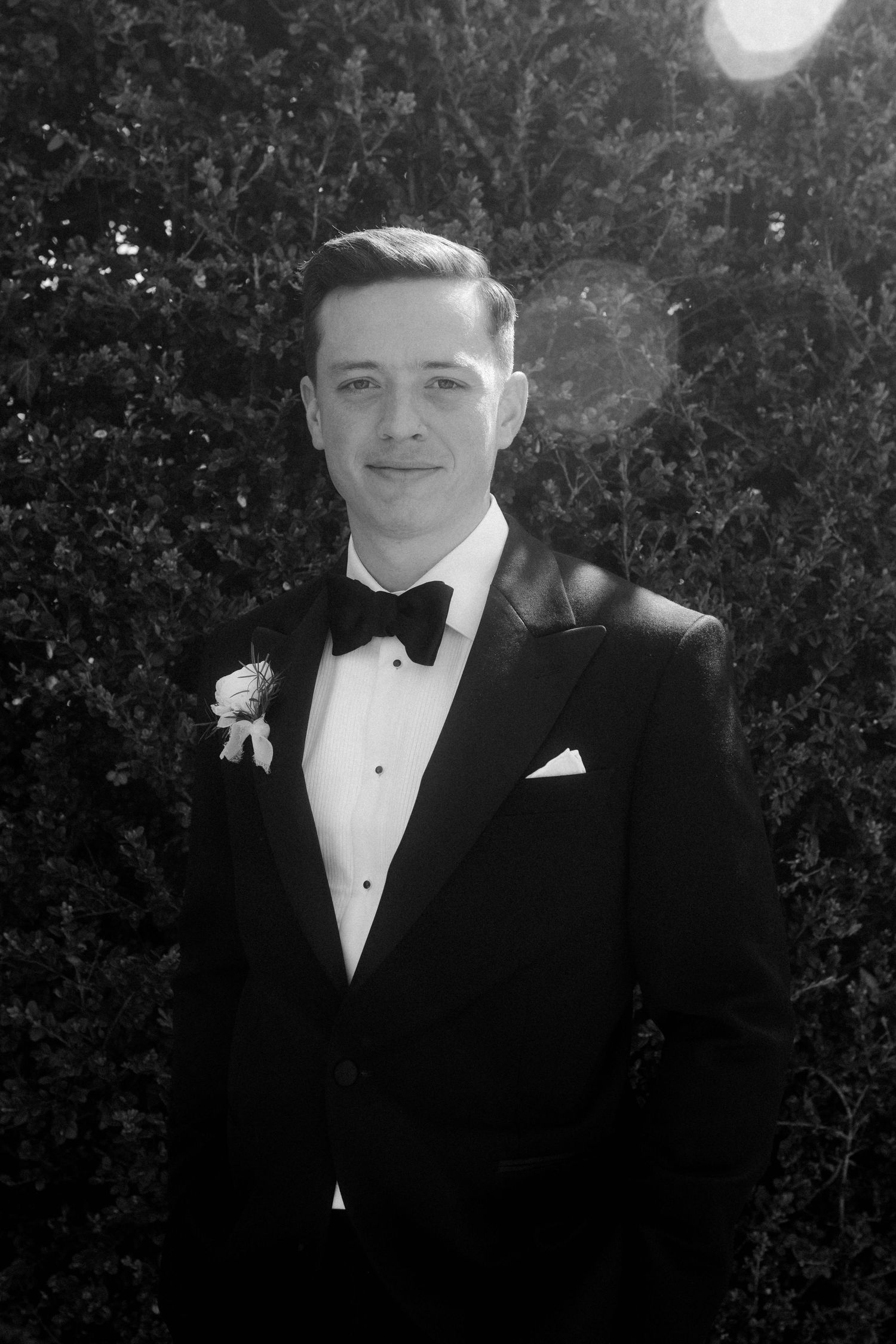 Black and white portrait of groom in tuxedo with boutonniere posing in front of lush hedges.