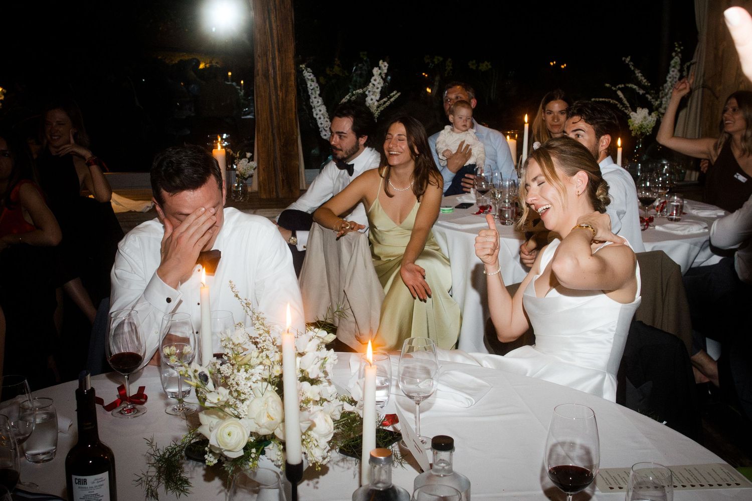 A bride and groom laugh at their wedding reception table surrounded by candles and white floral centerpieces at night.