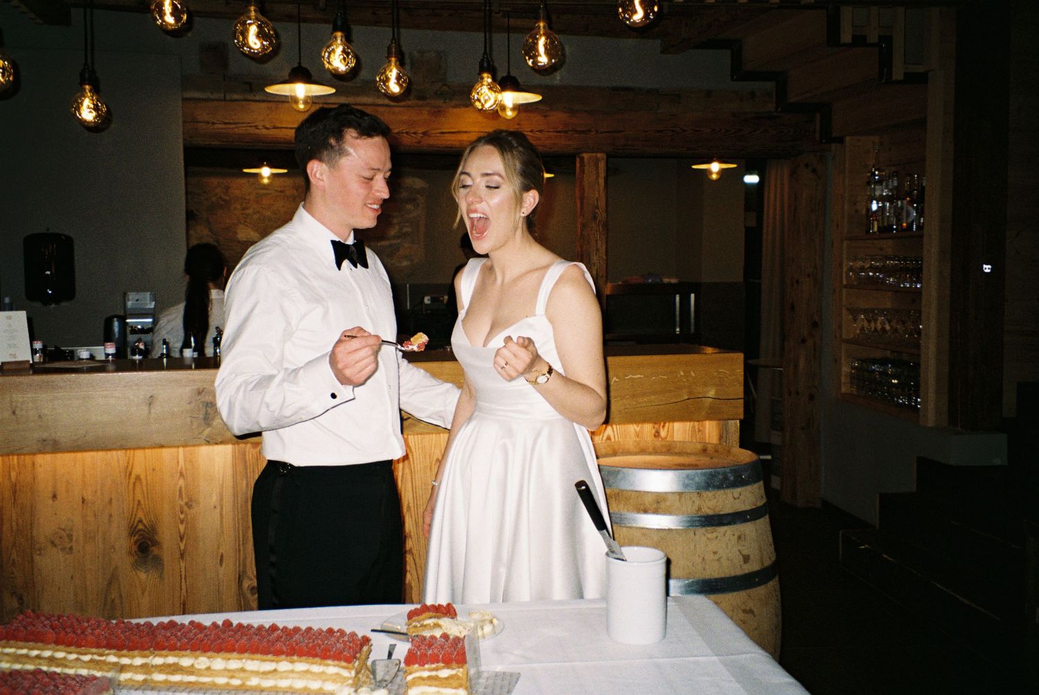 Bride and groom cutting wedding cake at rustic venue with warm Edison bulb lighting and wooden bar backdrop.