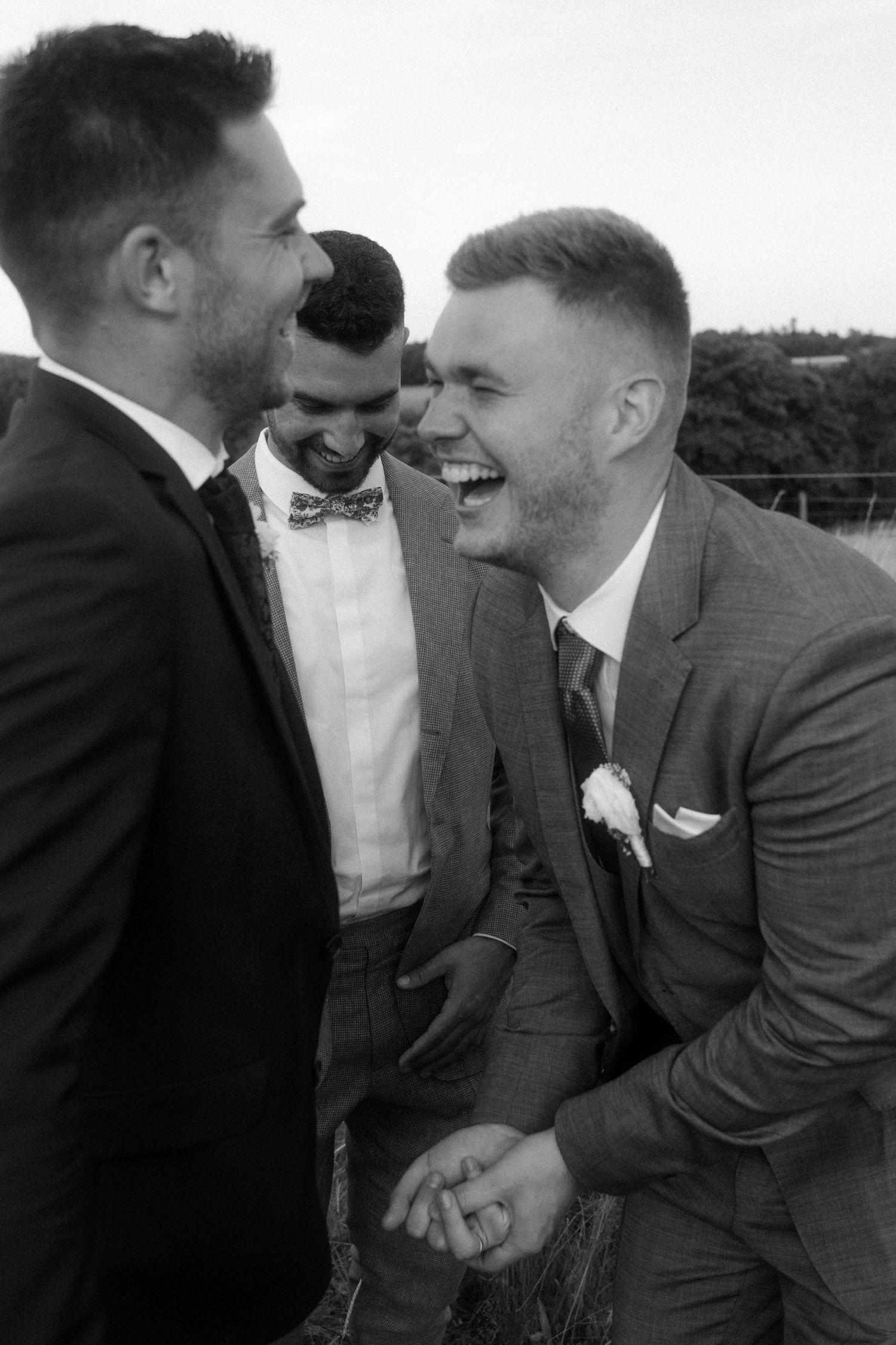 Black and white photo of two grooms laughing joyfully at their wedding, with a smiling groomsman in a bow tie behind them.