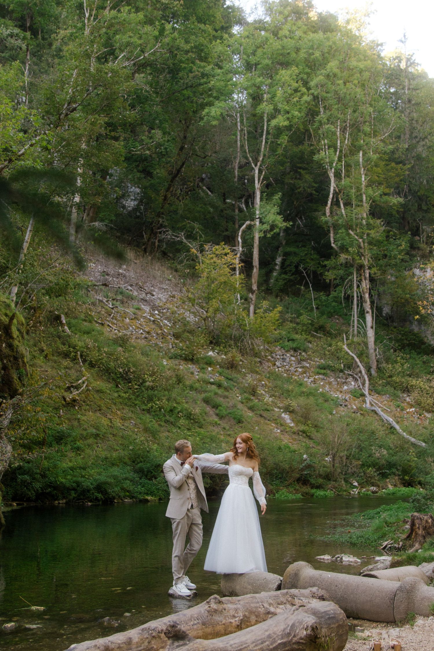 A bride and groom stand on rocks by a serene forest river, the groom helping balance his redheaded bride in a white gown.
