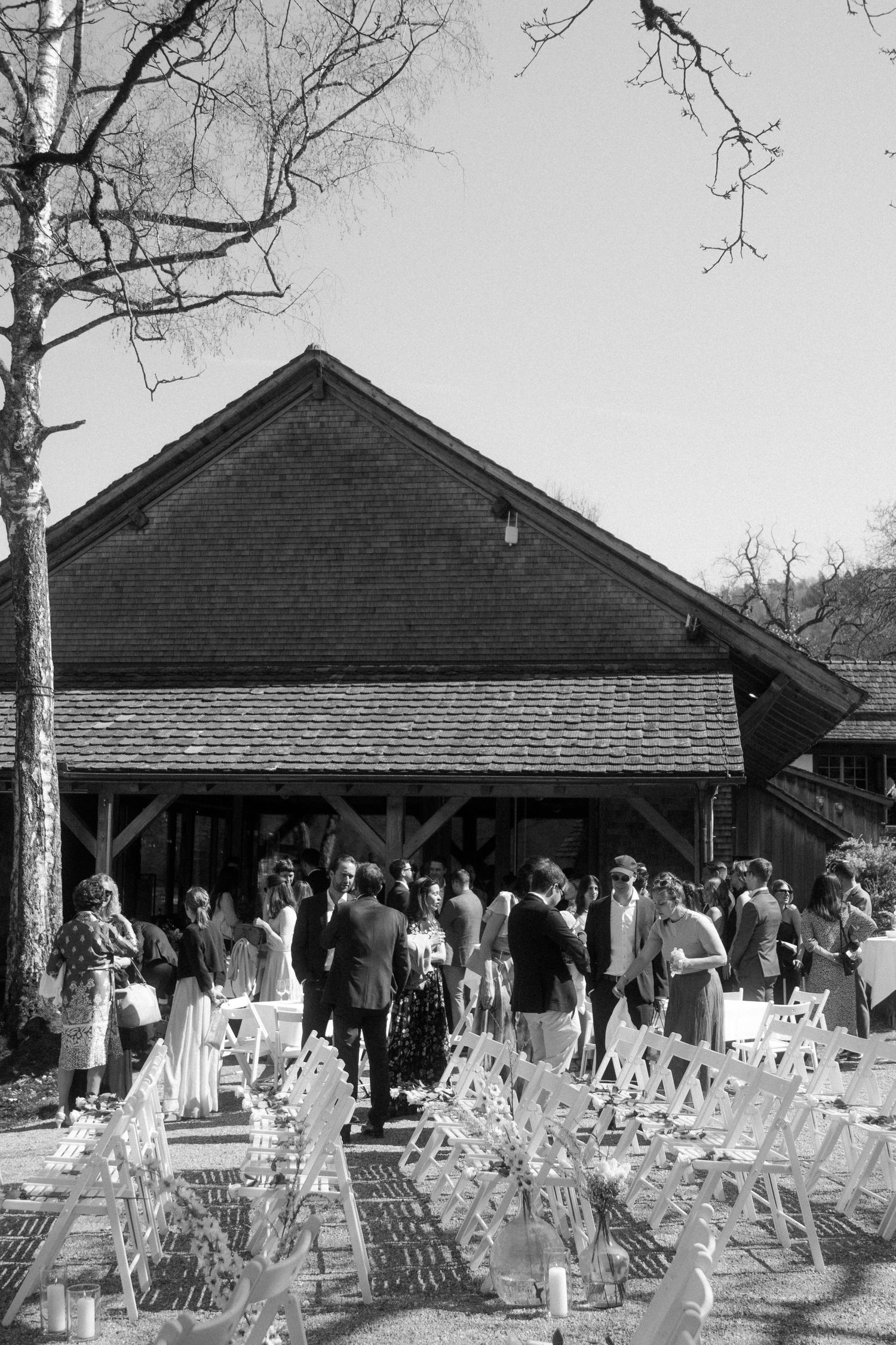Black and white photo of wedding guests mingling outside a rustic barn venue with white chairs arranged in rows.