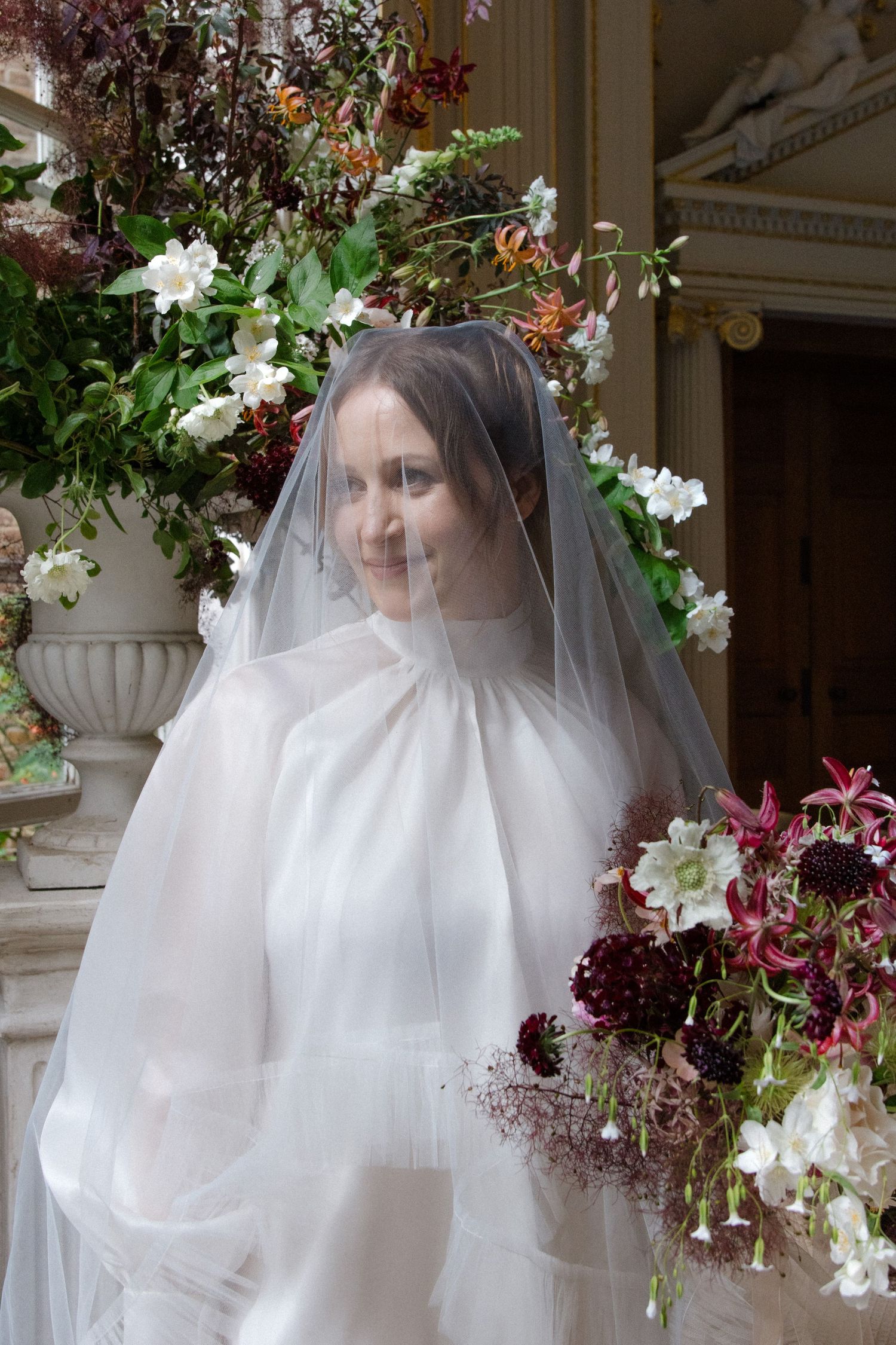 Smiling bride in white gown and veil holds burgundy floral bouquet beside ornate stone urn with lush floral arrangement.