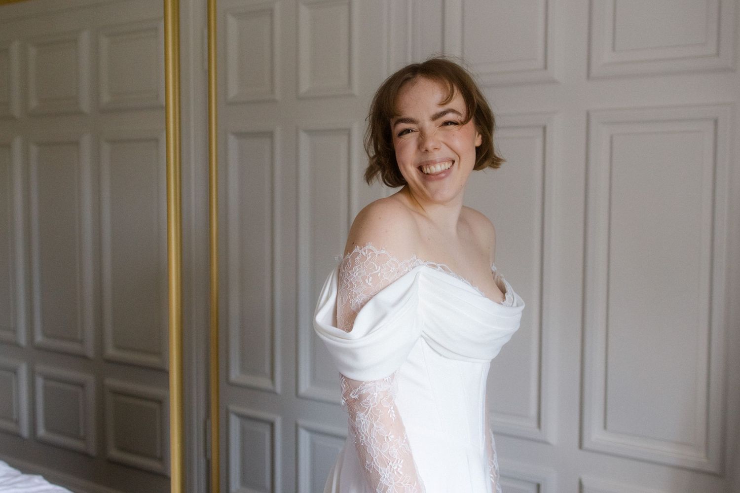 Smiling bride in off-shoulder white wedding dress with lace sleeves posing in elegant paneled room.
