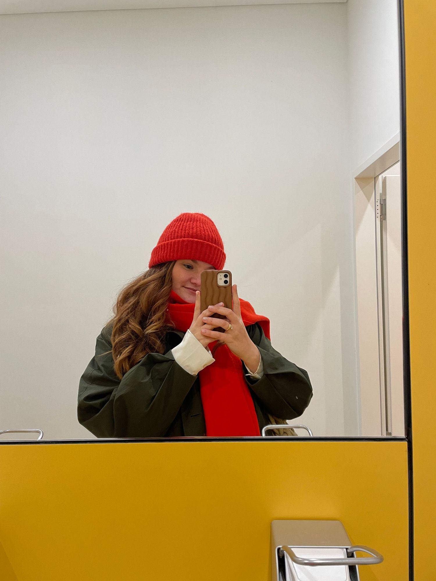 Person in red beanie and olive jacket taking mirror selfies against yellow and white wall panels.