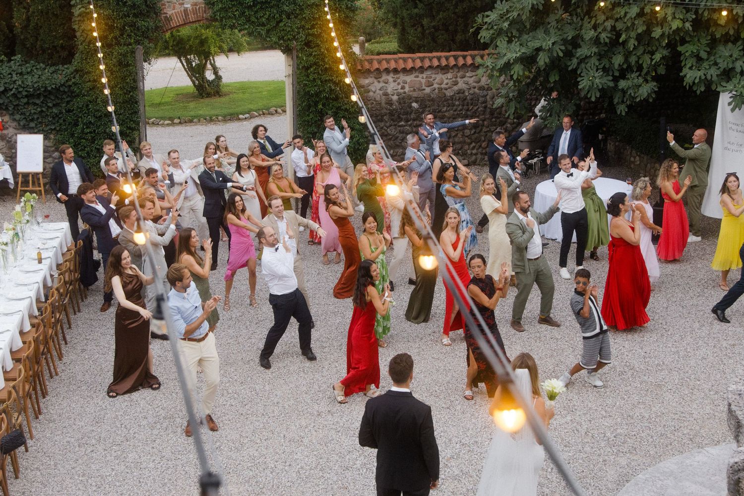 Wedding guests dancing in a lively outdoor celebration with string lights overhead.