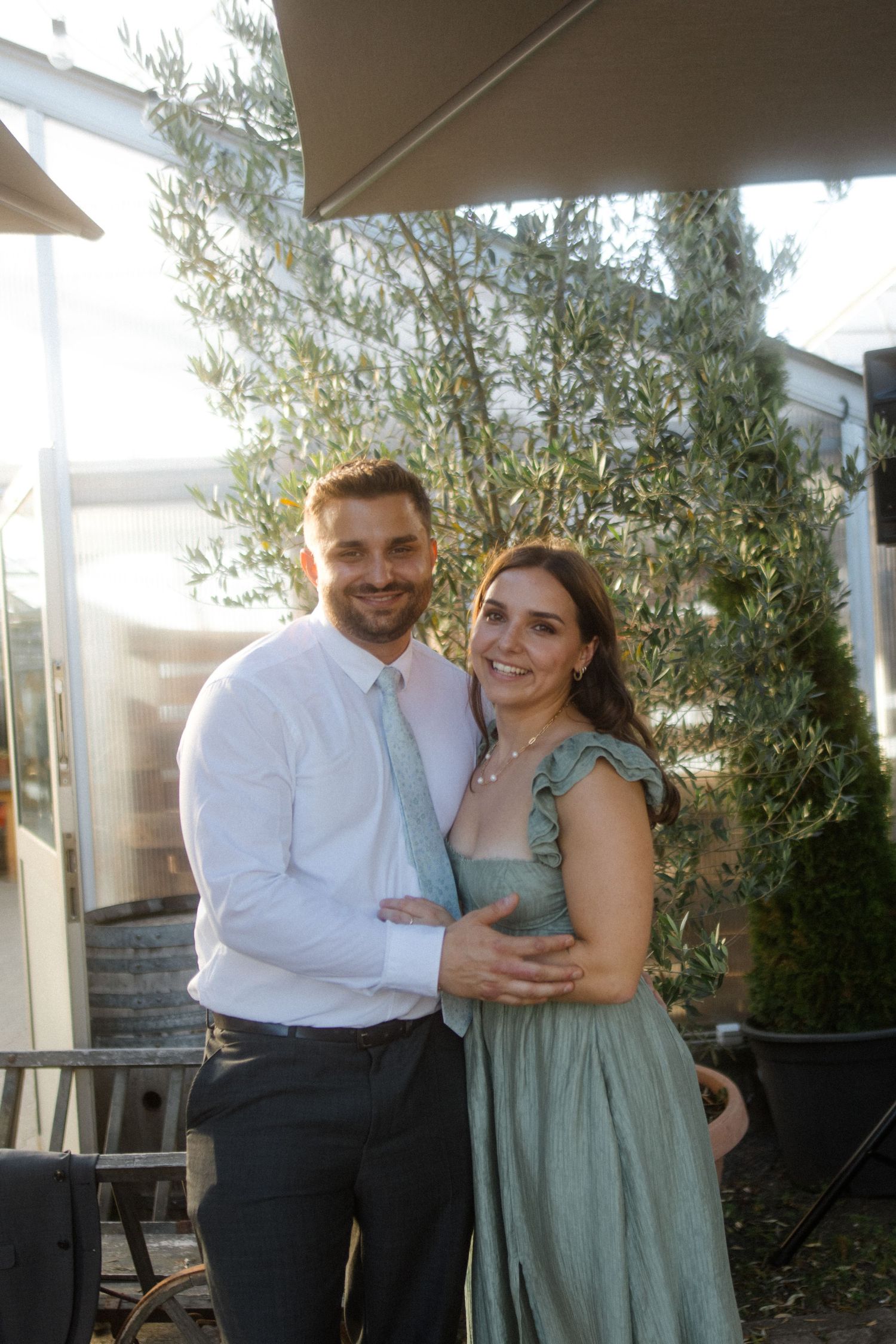 A couple poses together outdoors on a sunny day near olive trees.