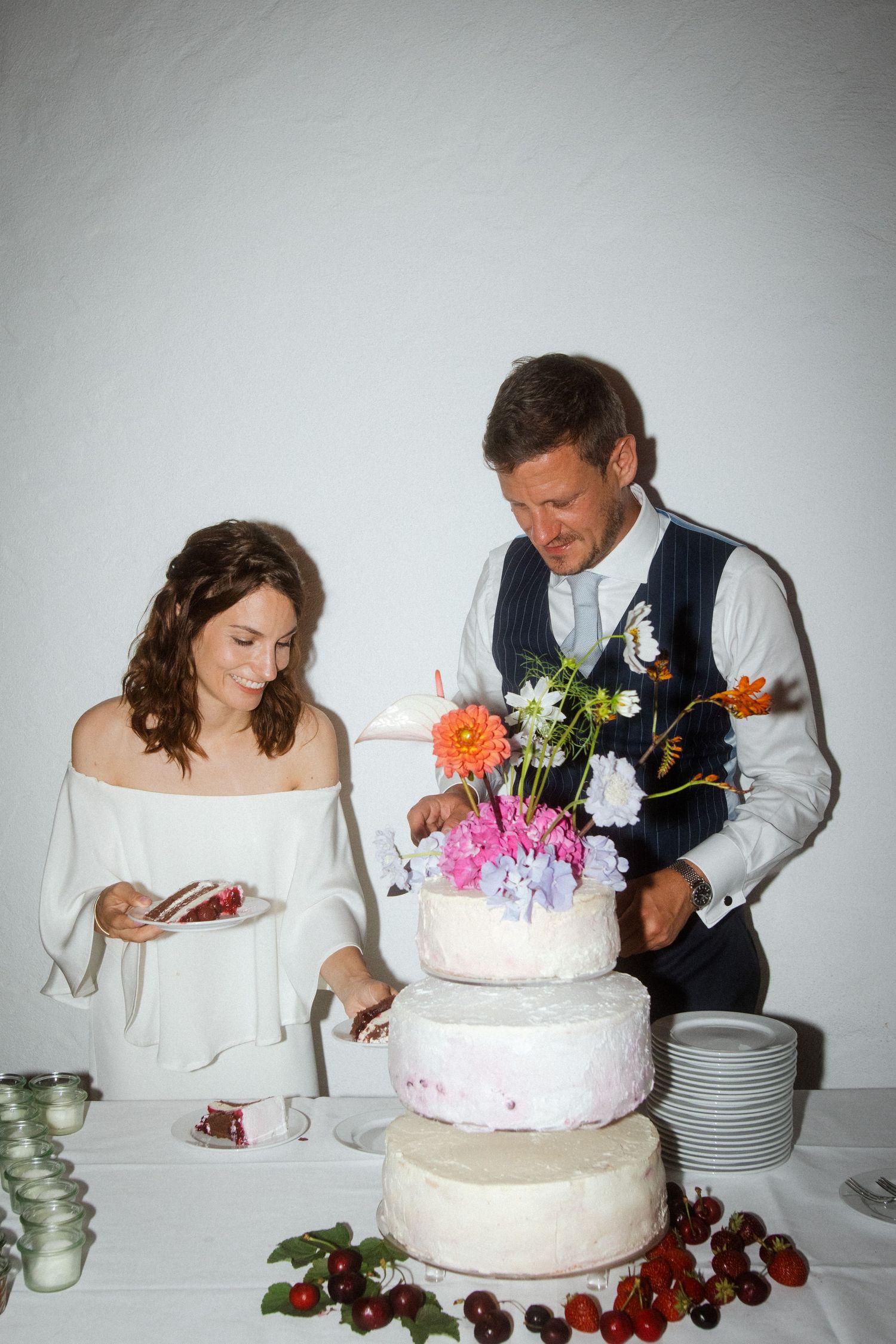 A couple cuts into their three-tiered wedding cake adorned with colorful fresh flowers.